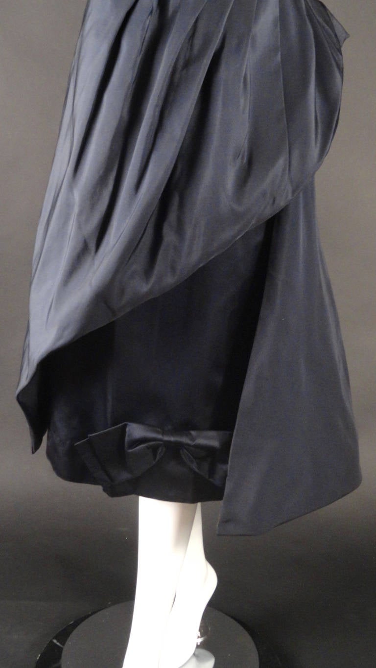Late 1950s dinner dress in a navy silk taffeta. The bodice has a scoop neckline with a center front slit. Dart fitted under the bust to the natural waistline seam. Slender skirt is dart fitted at the hips with a sweeping overskirt that falls in