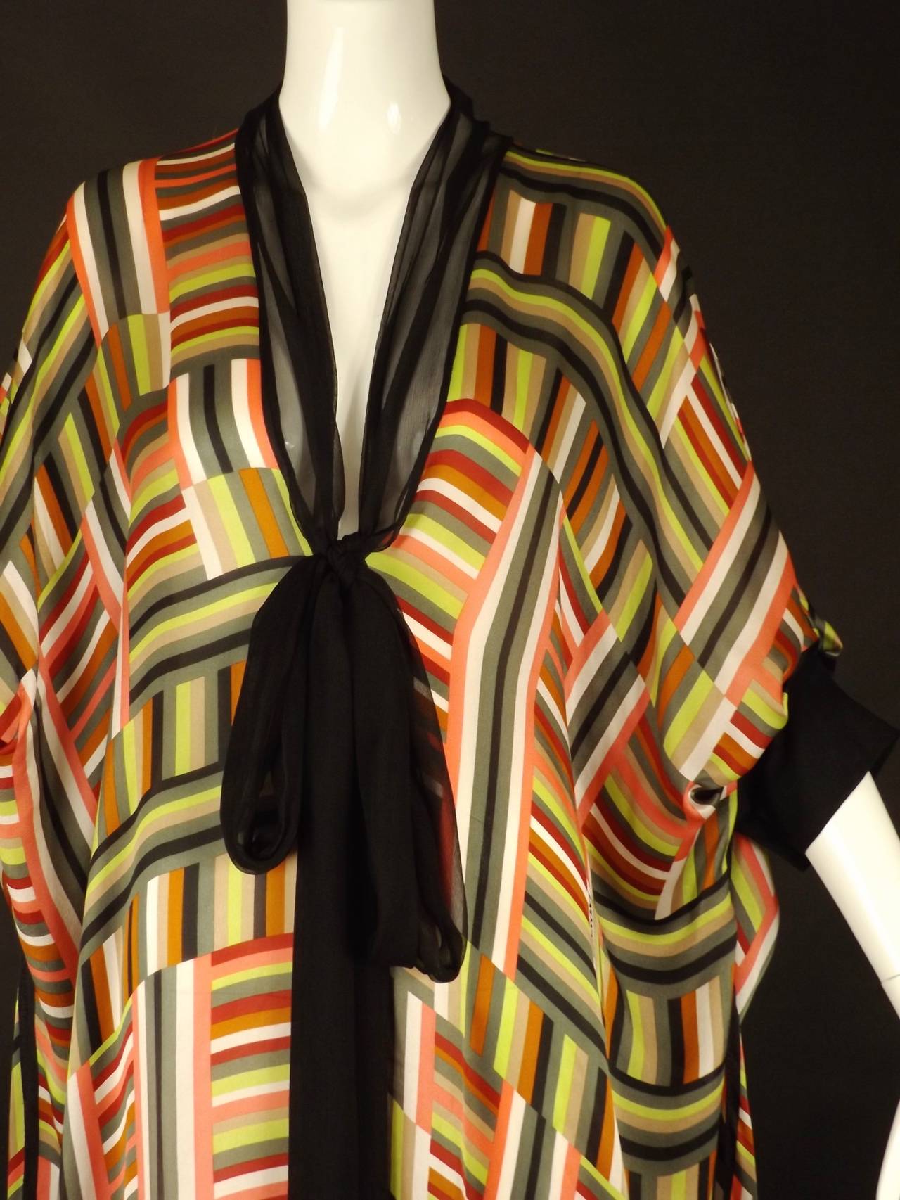 Awesome 1990s kaftan in a multi color stripe and geometric patterned silk chiffon. Plunging neckline edged in black chiffon with a long neck tie down the front. The upper sides are pleated into the black button closing cuffs. Slits down the side