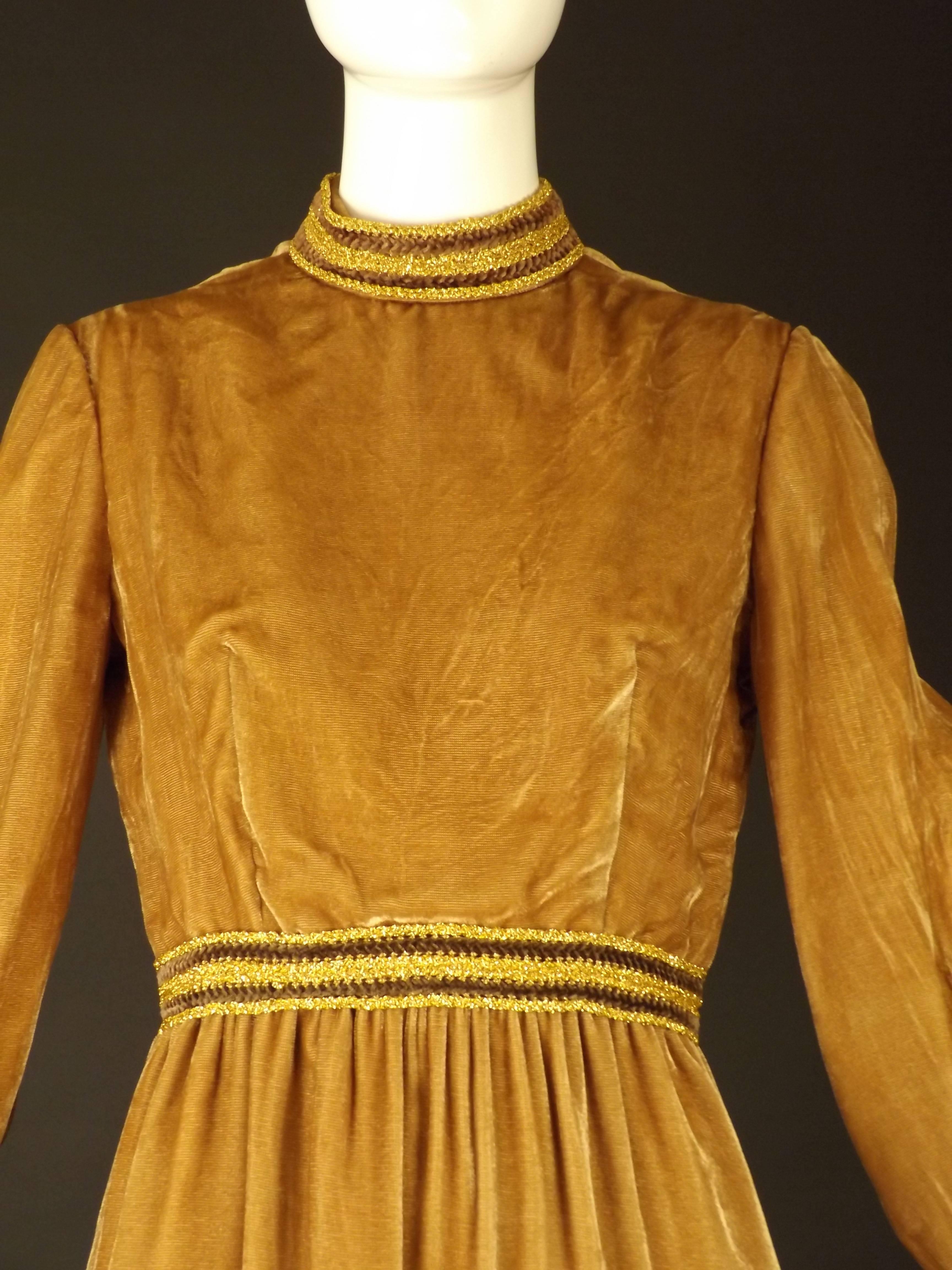 Late 1960s/early 1970s dinner dress in a camel colored textured velvet. The dress has a stand collar adorned in a gold lurex and brown chenille trimming. The bodice is dart fitted from the bust points to the natural waistline seam, that is also