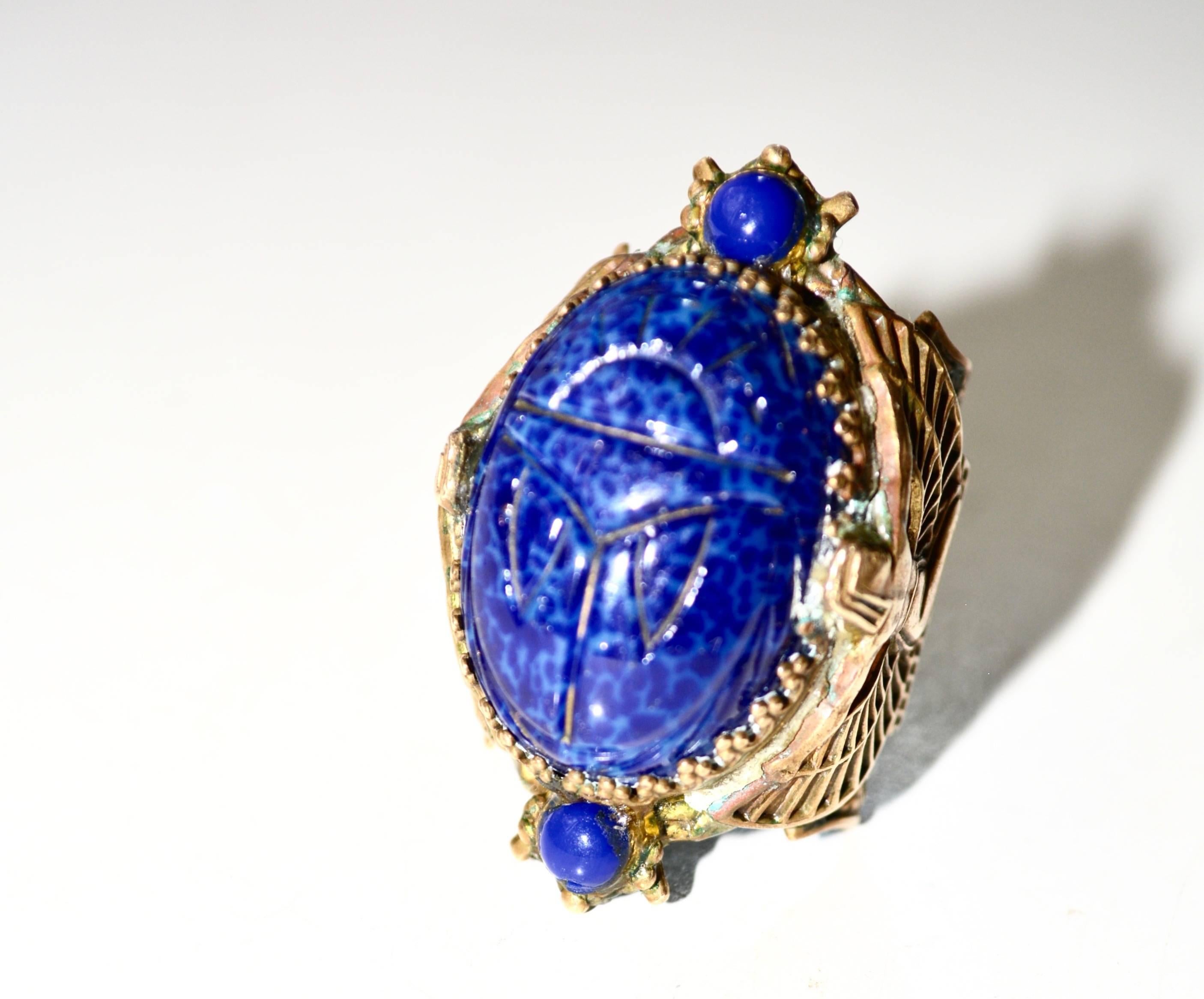 Brass and lapis colored glass scarab ring with Isis flanking the sides. Unsigned. Great example of the Egyptian revival style. Condition is very good with only minimal wear to one of the lower small glass stones and verdigris on the brass.  Somewhat