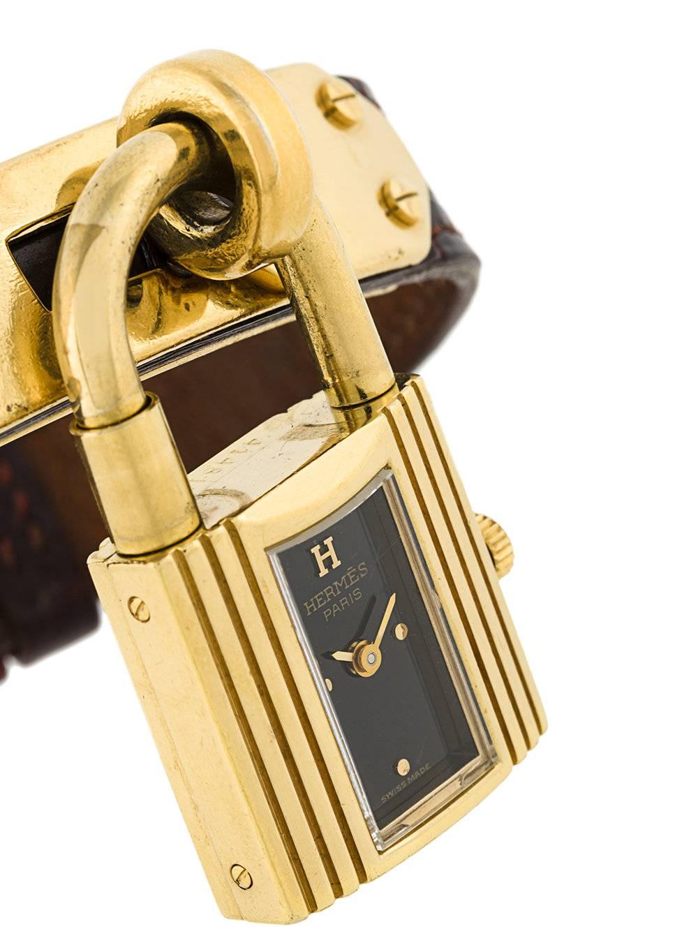 'Kelly' watch by Hermès in golden metal and bordeaux crocodile leather. The piece is hand wind and features two gold-tone hands with black background in a charming dangling padlock. The item was produced in 1989 and is vintage, it comes in very good