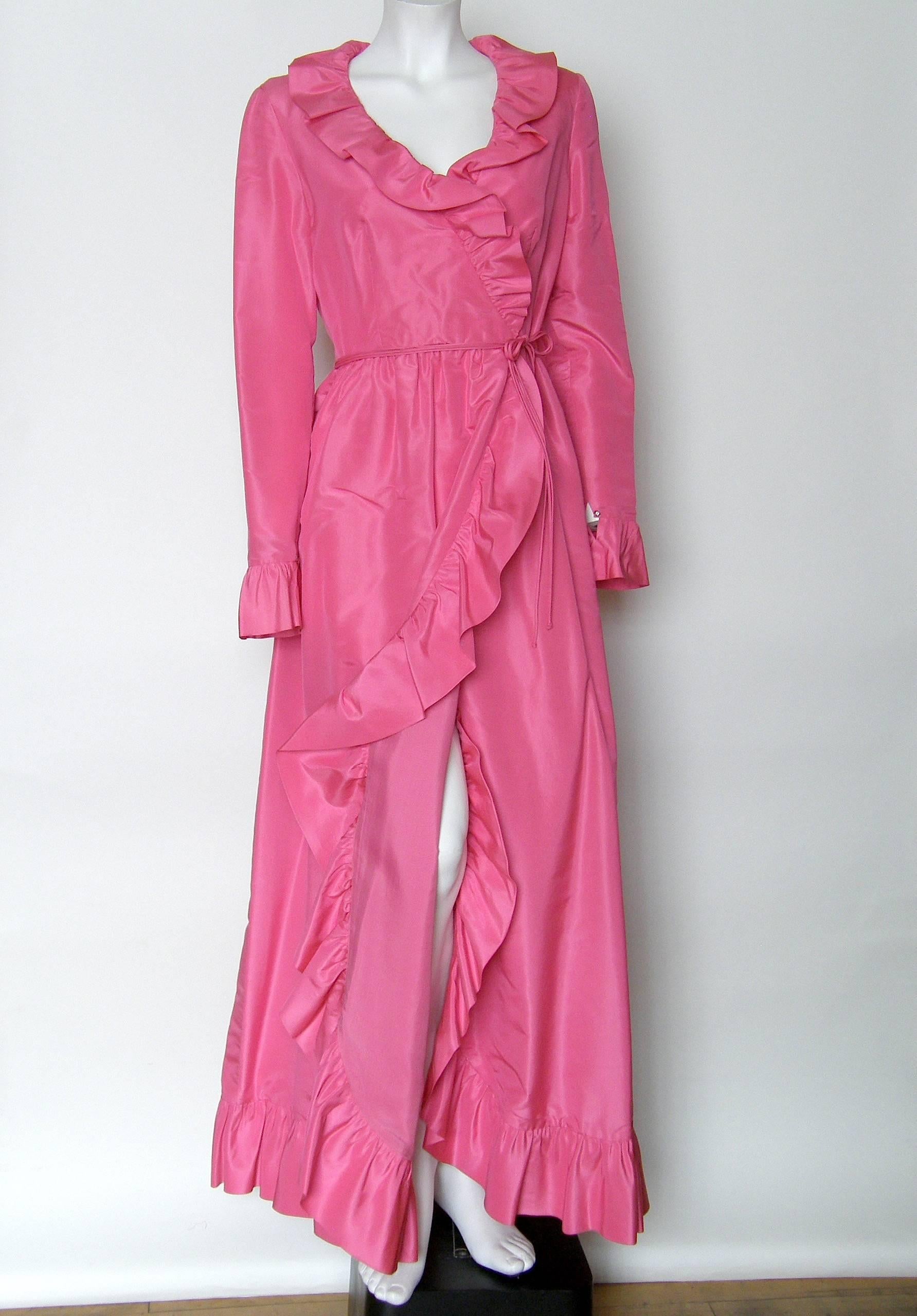 This Mollie Parnis pink taffeta gown has a fixed wrap style with a sexy overlap/slit in front for a peak of leg. The edges are trimmed with playful ruffles, and the fitted sleeves have ruffled cuffs. The attached double cords tie at the