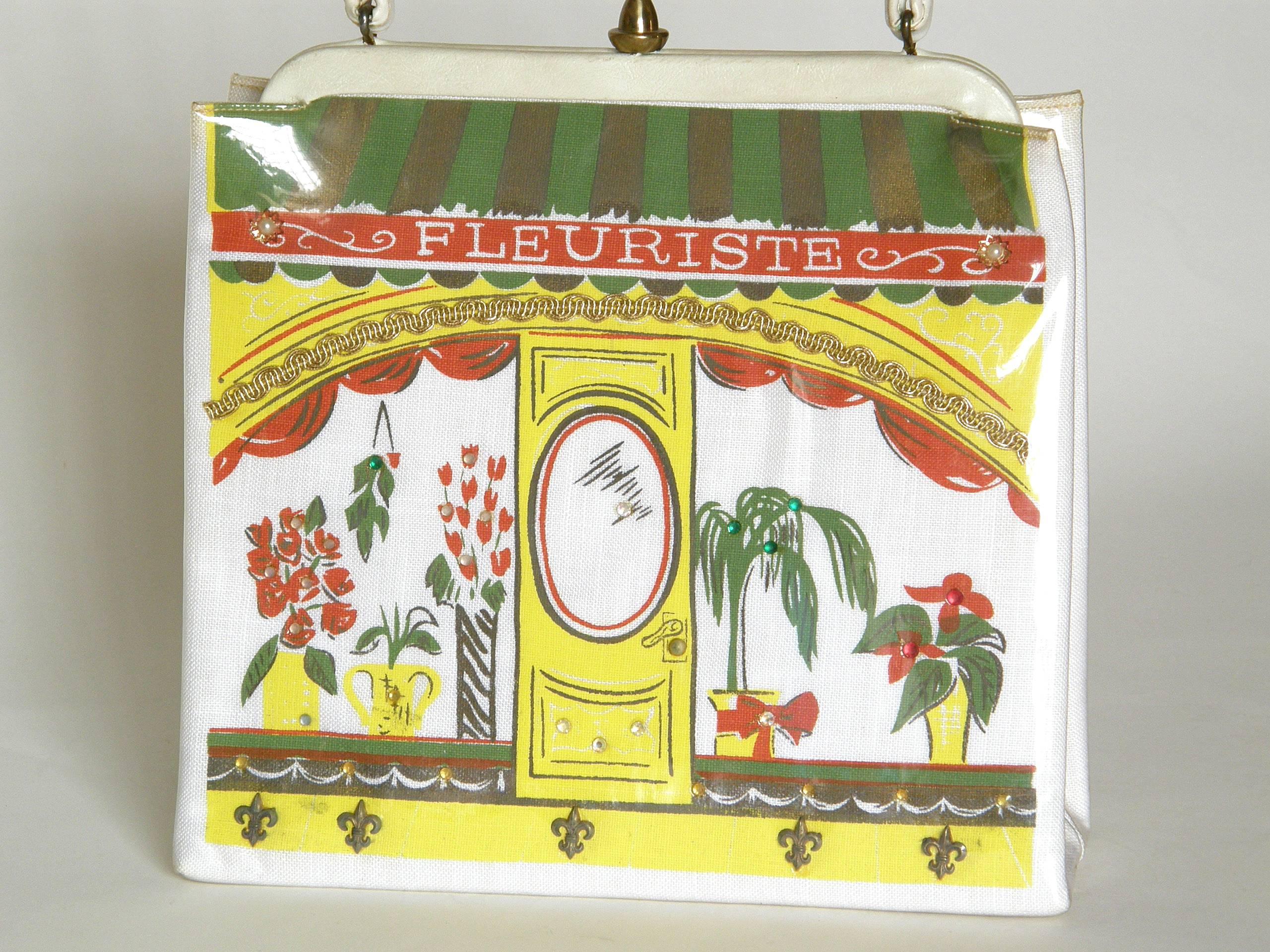 This charming, novelty handbag by Soure' is designed like a French florist shop. The front of the bag has a screen printed image of a storefront with flowers and plants in the display windows that flank the front door. The striped awning has the