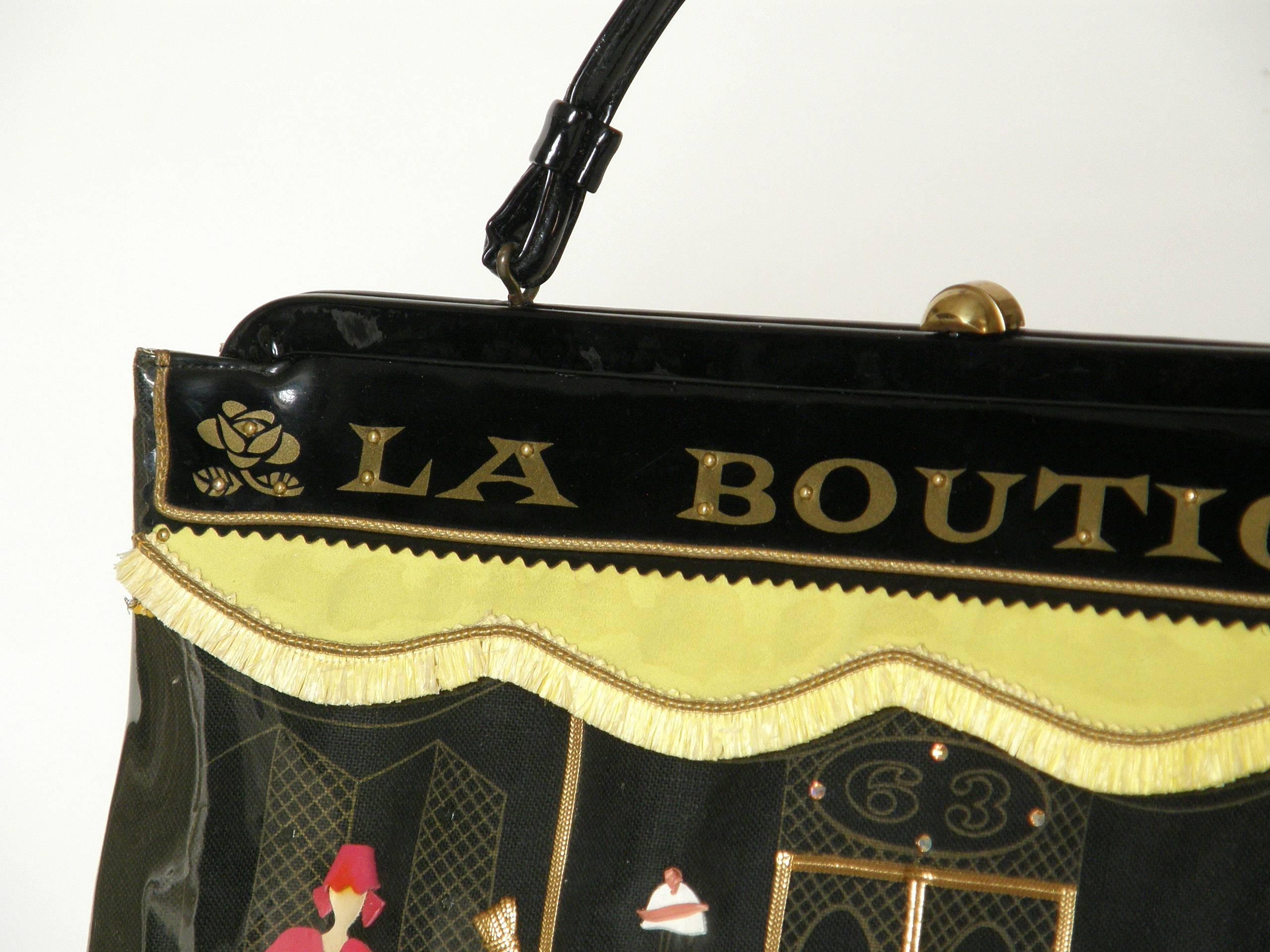 This stylish, novelty purse by Soure' is designed to look like a French clothing shop. The front of the bag has a screen printed image of a storefront with mannequins, clothing and accessories in the display windows that flank the front door. The