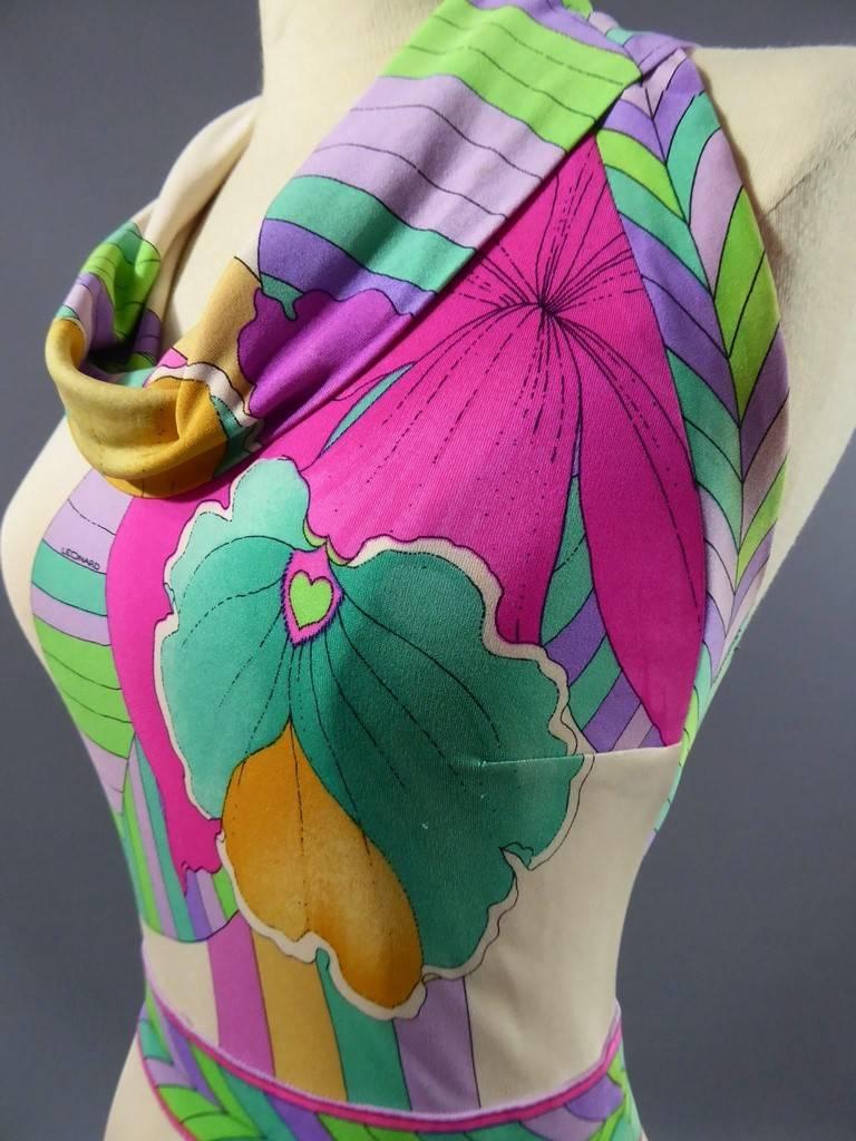 Circa 1990.

France.

Leonard dress in printed jersey. Sleeveless, backless, back closure with a pink zip. Beige background and large patterns of exotic pink, green, turquoise, purple and yellow flowers. Trompe l'oeil waist belt with geometric