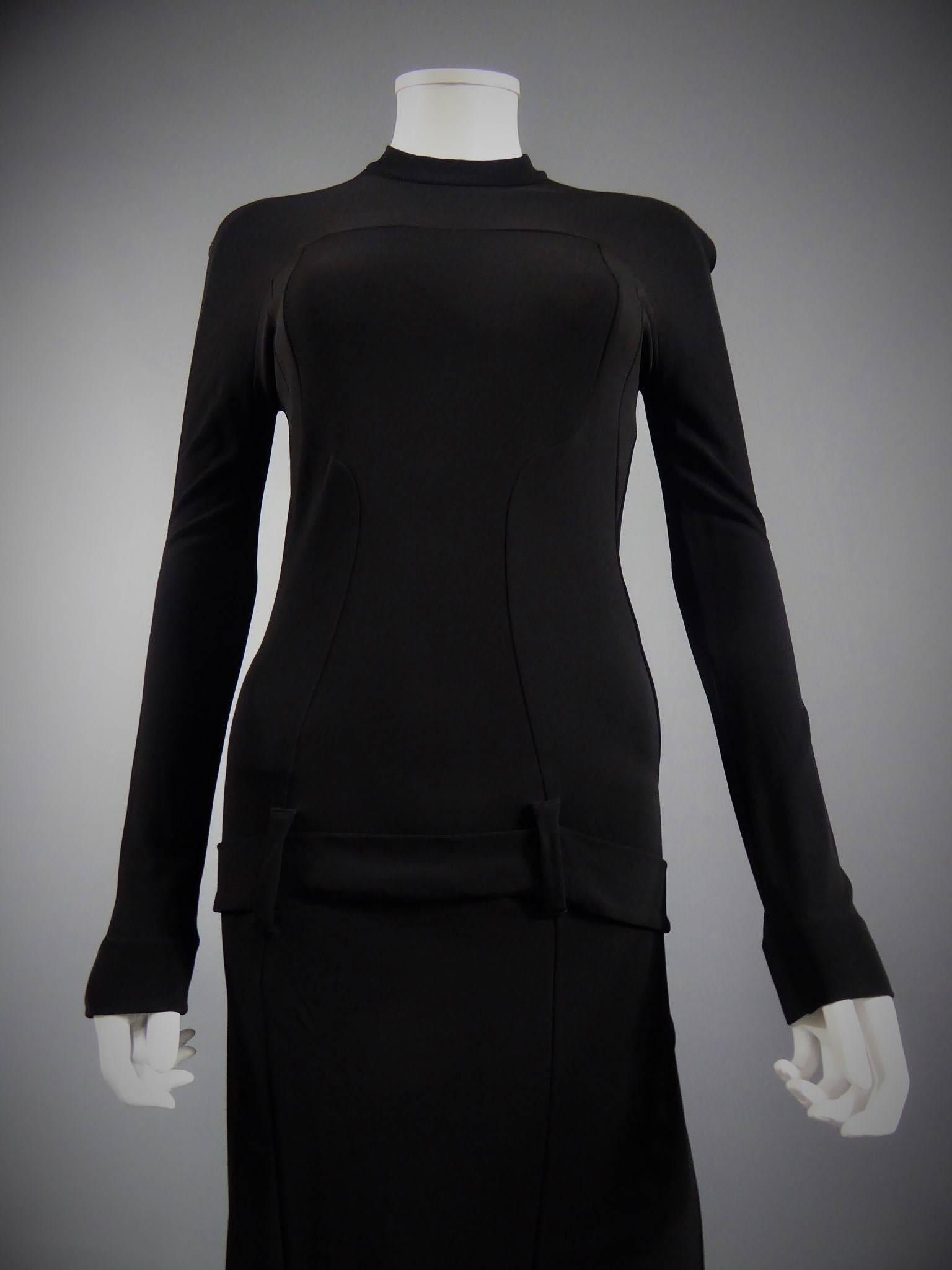 Circa 2000

France

Balenciaga black dress in elastic black jersey. Long sleeves. Nicolas Ghesquière line. Details on the sides, half belt on the front ending at the buttocks. Mid-long skirt under the dress. Zip closure in the back. Very fitted.