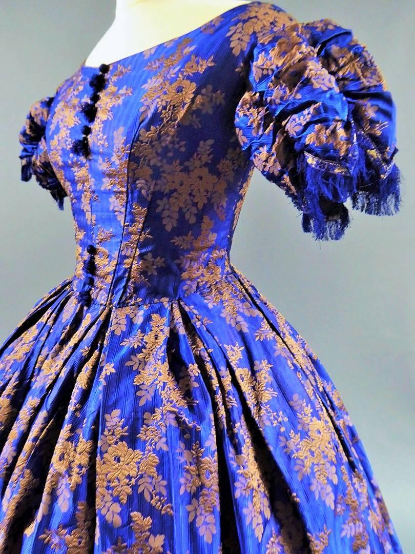 Circa 1850
England

A crinoline ball-gown in a rich deep blue façonné striped silk, adorned with brocaded tobacco and copper flowered stems. Whalebone bodice is assembled to the skirt with flat pleats . The gown is entirely lined with a green glazed