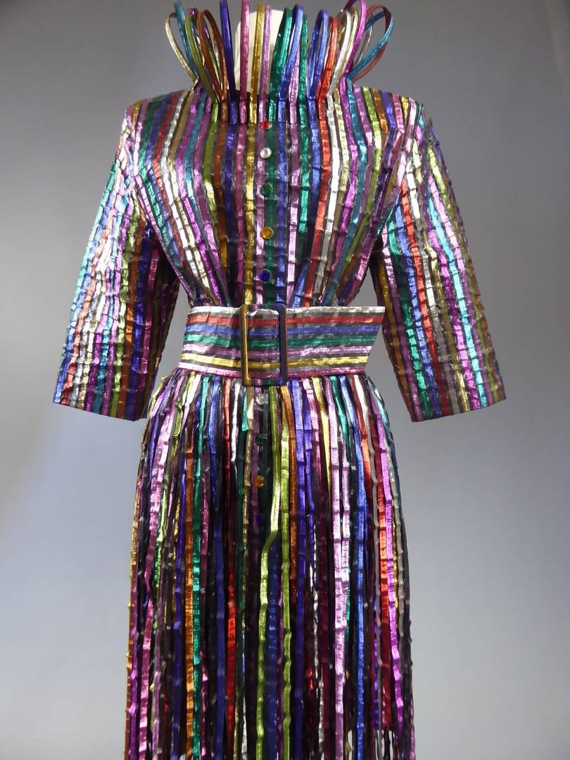 Circa 1992.

France.

Ensemble comprising a long jacket and skirt belted with multicolored iridescent strips looking like the colors of the rainbow. Rigid laminated jacket in similar strips. Button front with multicolored rhinestones. Large and
