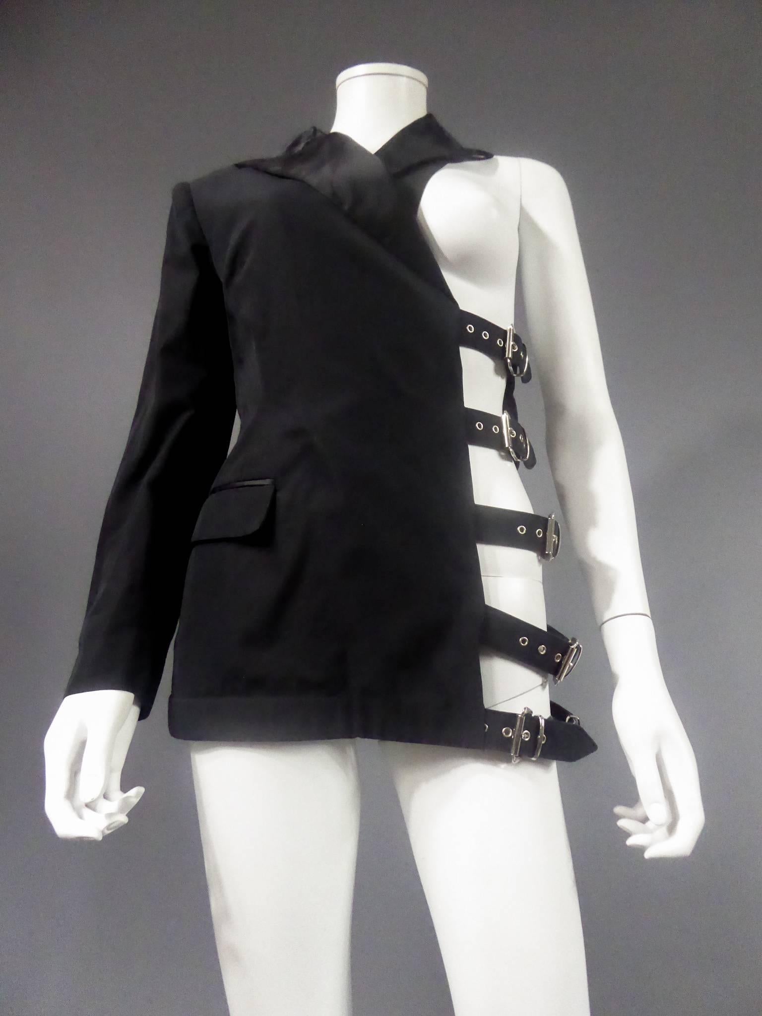 Circa 1980

France

Fetish inspired asymmetrical tuxedo jacket in cotton and wool gabardine. Black satin collar with recall at the pockets. Rigid leg at the bottom of the jacket for support. Lining black leather legs, buckles and rivets in gray