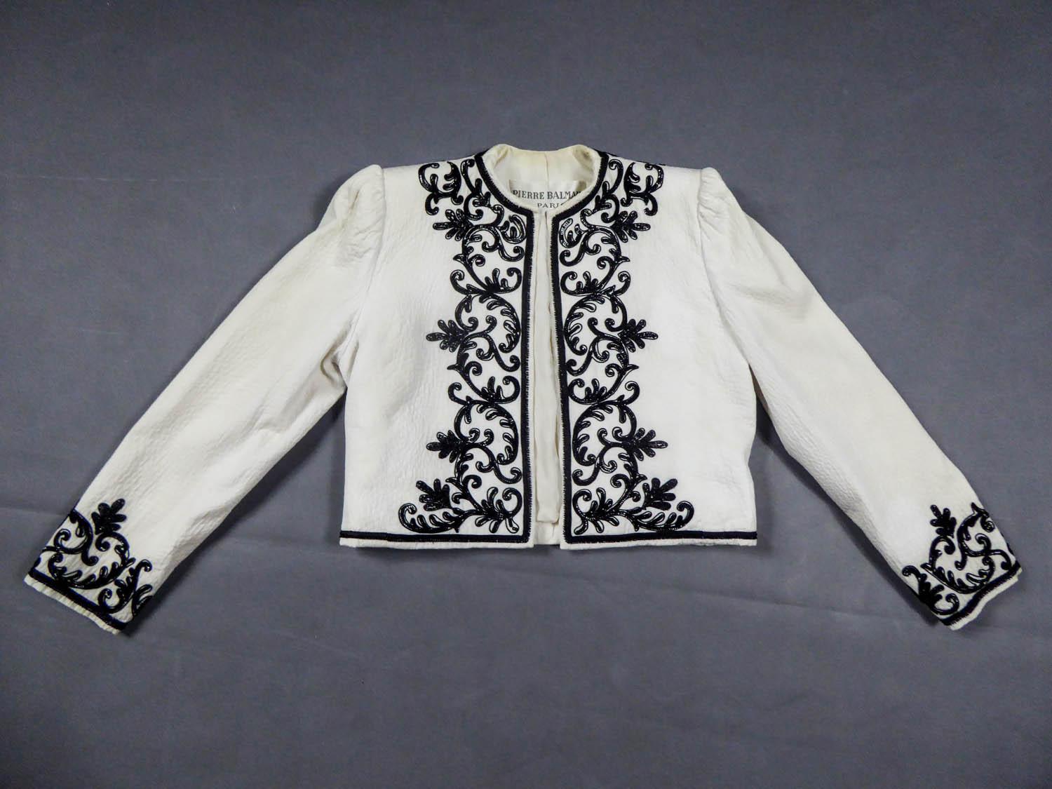 Circa 1955/1965
France

Elegant Pierre Balmain bolero in embroidered white cotton piqué with round collar and small epaulets, dating from the late 1950s. On the edge of jacket and wrists, very fine work of lying black sequins embroideries set with
