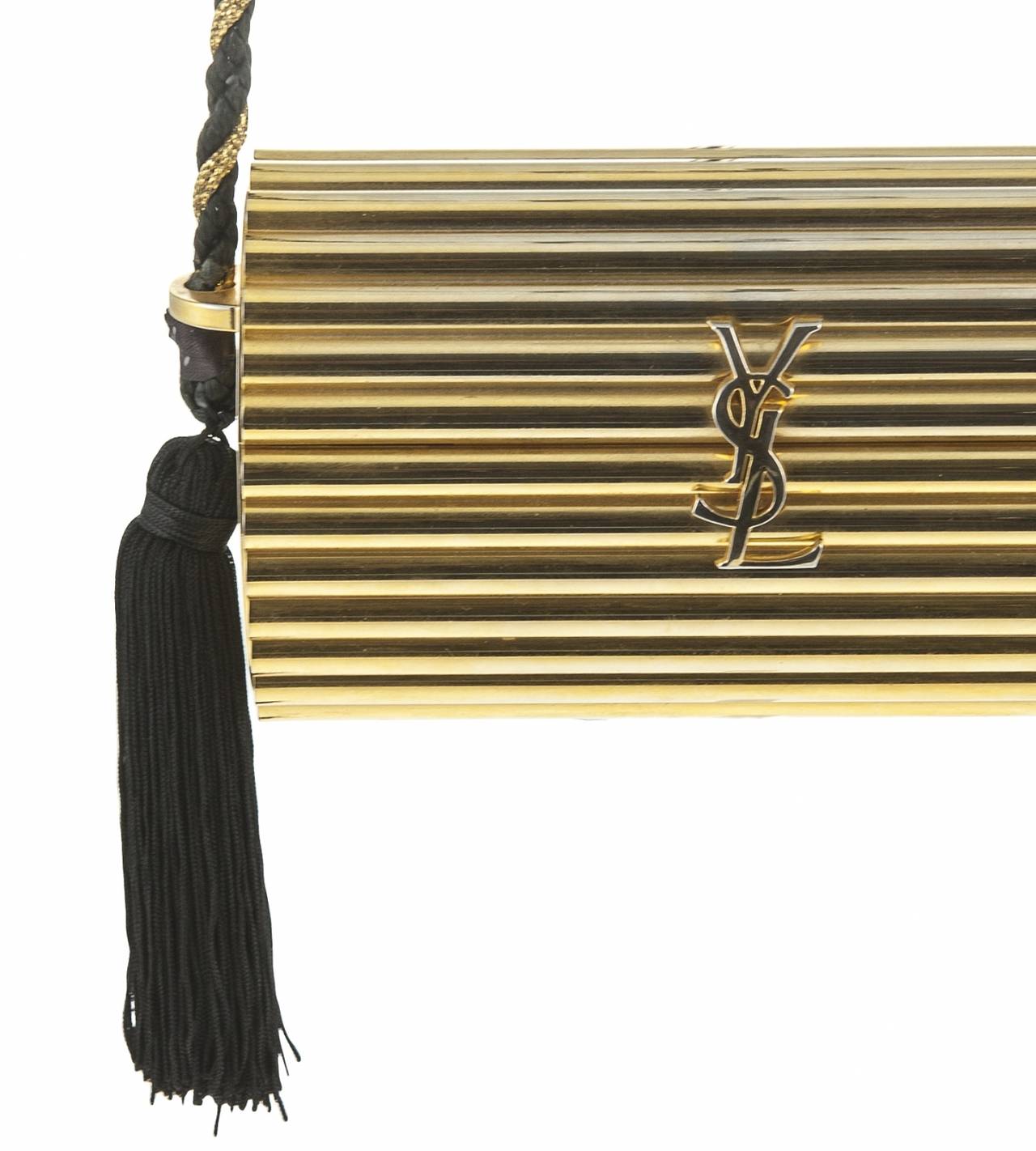 Vintage Yves Saint Laurent Gold Hard Case, Tube Shape, Evening Bag With Black And Gold Cord Strap With Hanging Tassels. YSL Logo Clasp.
Width - 15cms
Height - 9cms
Depth - 5cms
Strap Length - 56cms
Excellent Vintage Condition.