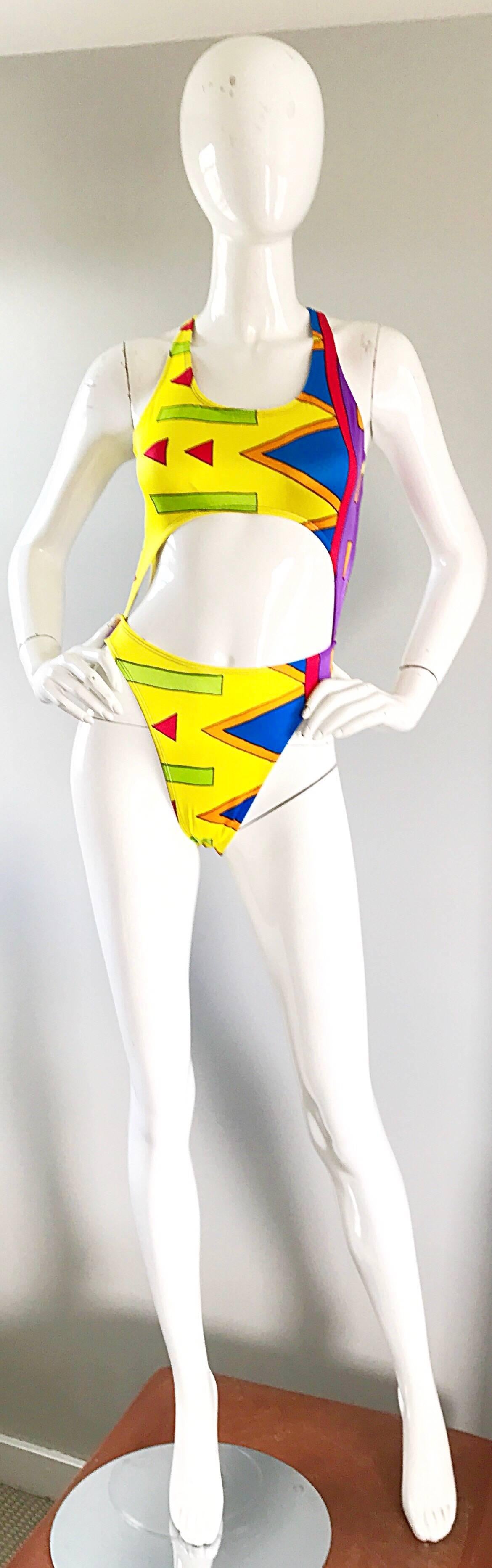 Sexy brightly colored early 90s cut-out one piece swimsuit or bodysuit! Features geometric shapes in royal blue, purple, red, orange and neon green throughout, on a bright yellow background. Straps criss-cross in the back. Cut-out detail at the