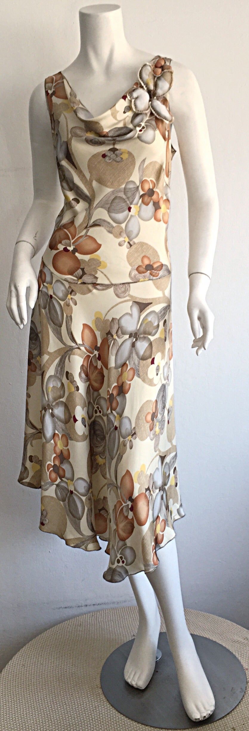 Pretty silk dress, by Rozae Nichols! Gorgeous neutral hues, with a unique floral print throughout. Flattering silhouette, with silk floral puff appliqués at left shoulder. Asymmetrical hem that 'flows' when on! Looks great alone, or with a belt! Can