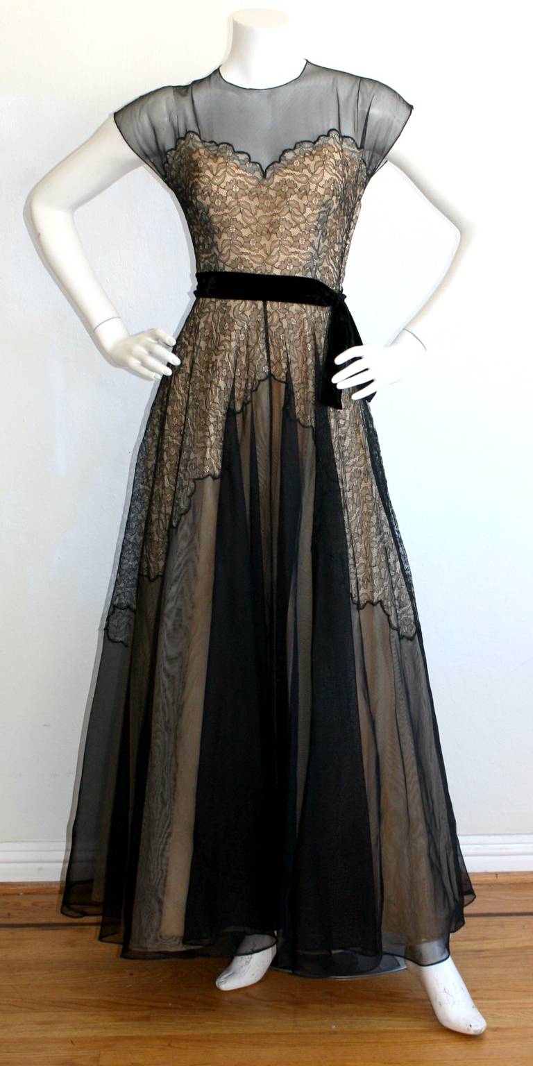 Featuring an incredible vintage 1950s vintage lace illusion gown. Great full skirt, with intricate black lace detailing under nude layers. Attached velvet bow belt, with hook and eye closure at side. Hidden zipper also up side. Approximately Size