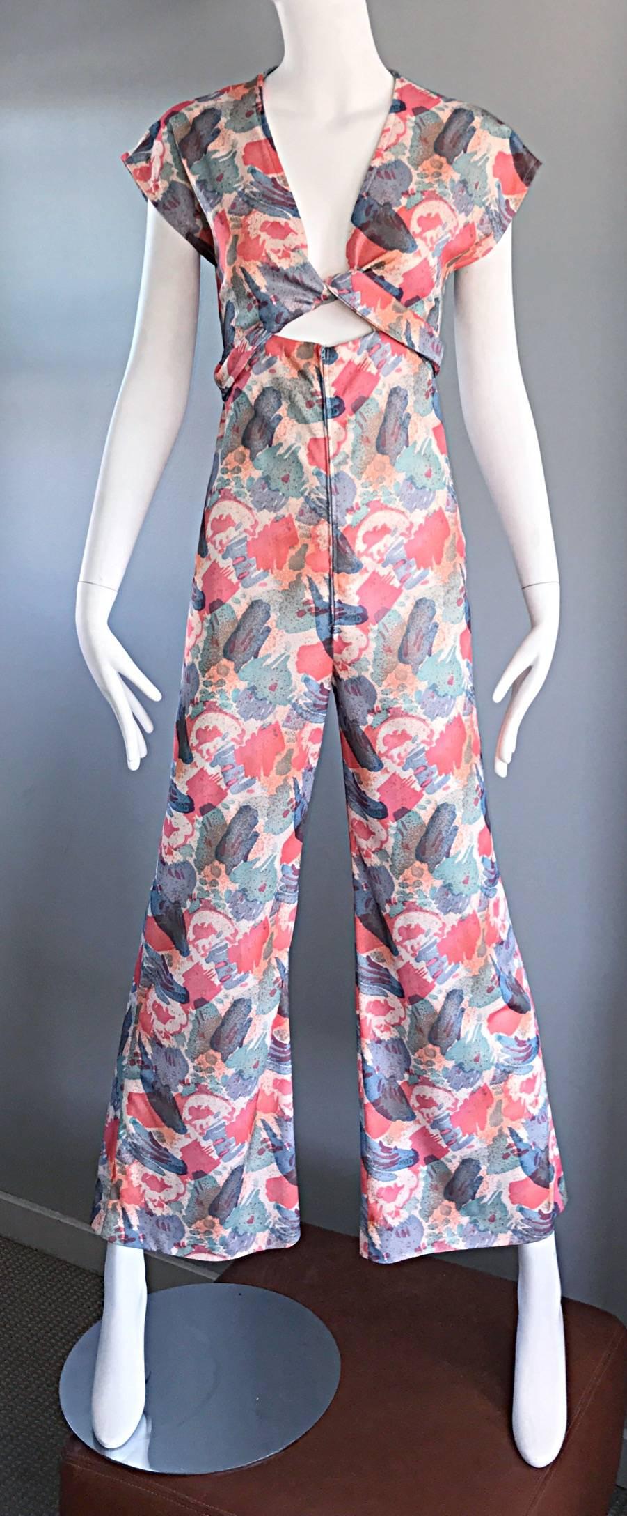 Smashing 1970s abstract cut-out wrap jumpsuit! Features warm hues of pinks, blues, greens, peach and ivory. Flattering cap sleeve, with a. Fitted bodice. Wide flared palazzo pants. Top ties in the back, and can be worn multiple ways. Looks chic with