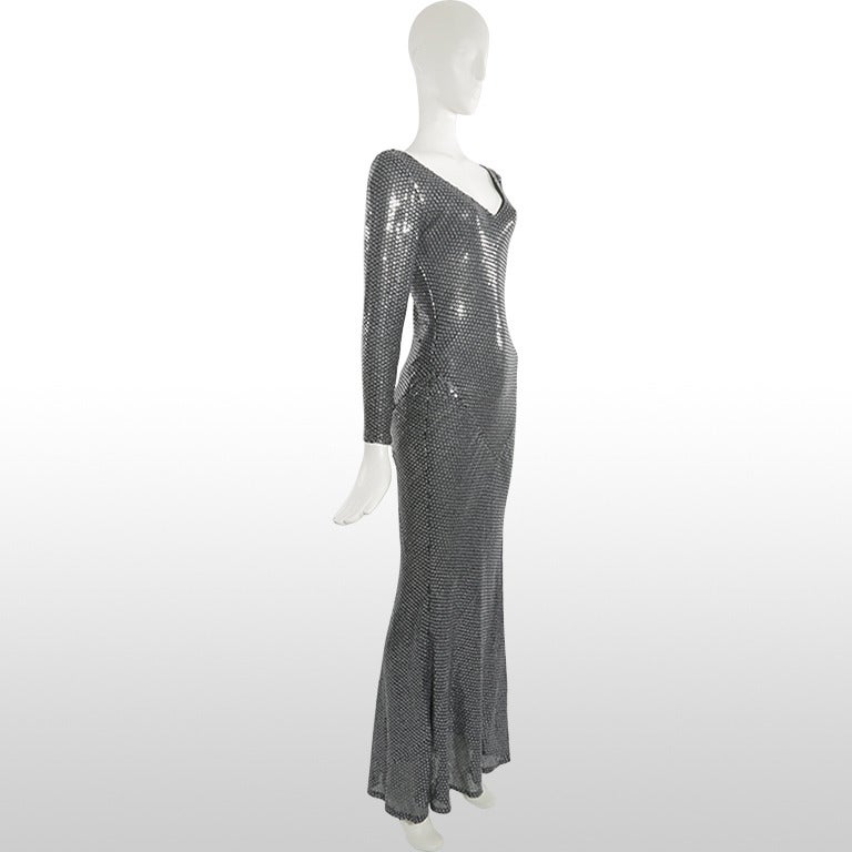 This elegant gown by Bruce Oldfield is a slinky number covered in printed sequins that create a shimmering steel grey effect. It has full length sleeves with zipped cuffs and a feminine plunging V neckline. The fabric is form fitting as it has