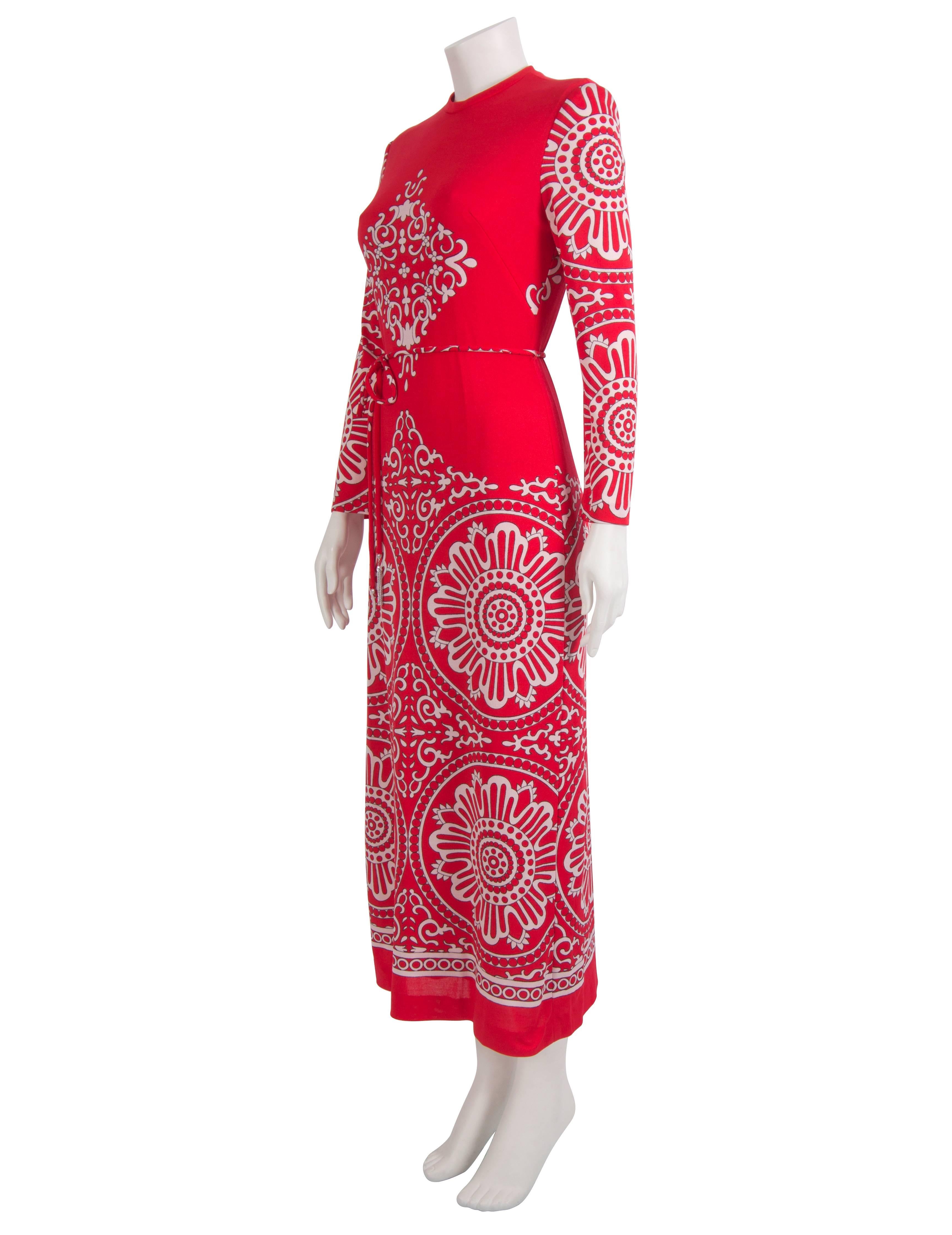 A 1970's red jersey maxi dress featuring a mirrored mandala print throughout. The dress closes with a zip at the centre back and is fully lined with a red mesh fabric. The dress features a belt with metal tassels (one missing - pictured). Excellent