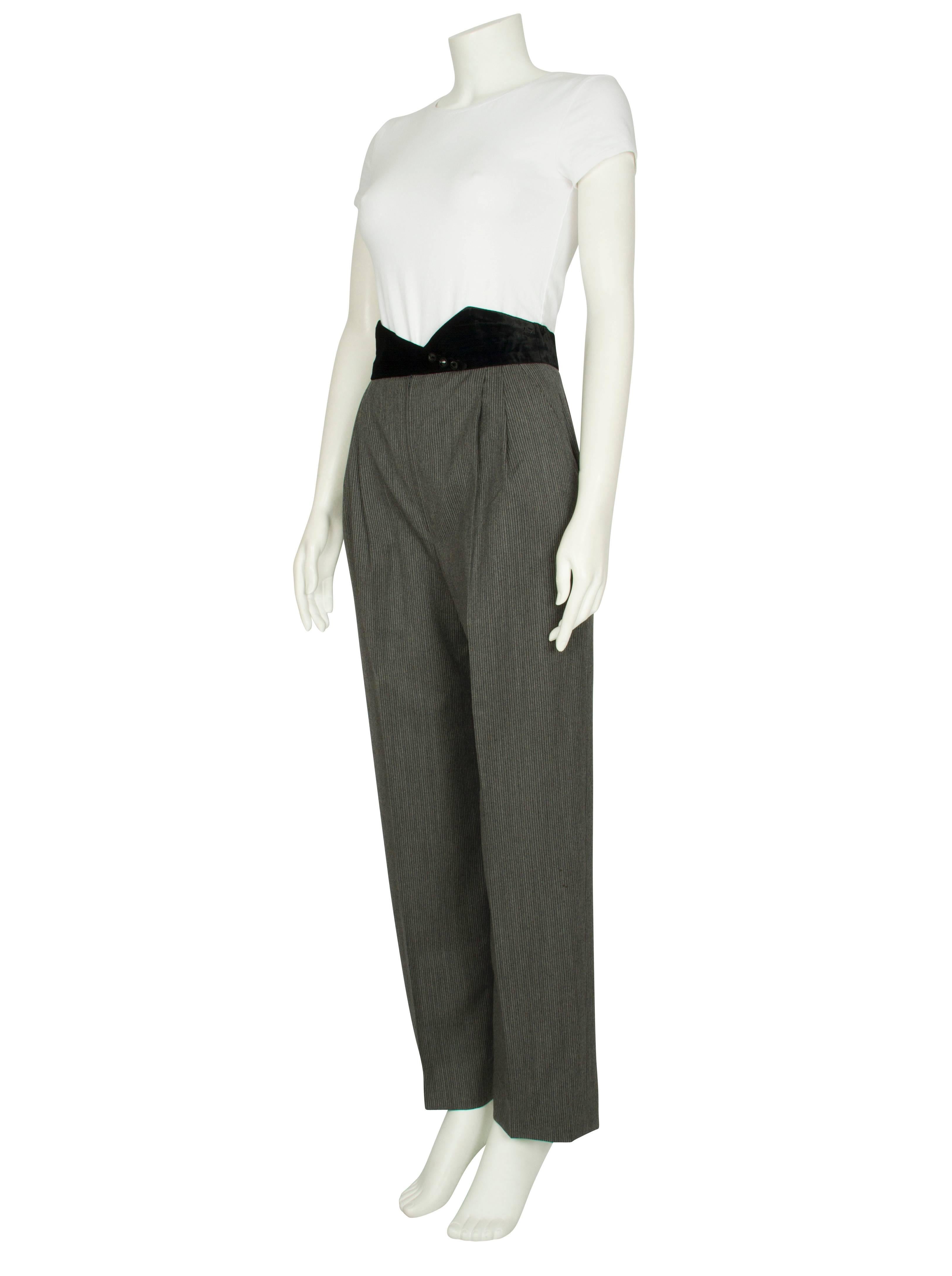 A pair of 1980s Ungaro Couture grey pinstripe wool trousers featuring a pleated front and an asymmetrical pointed velvet waistband embellished with jewelled buttons. The trousers have knife pockets at the hips and tapered legs. Excellent vintage