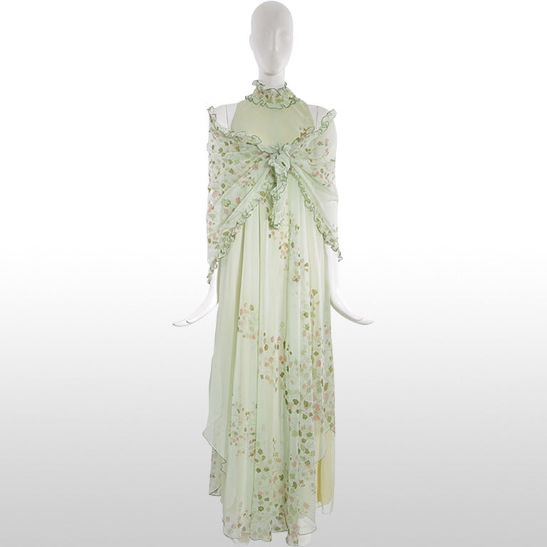 This gorgeous 1970's gown is made from a mint green chiffon overlay printed with an autumnal foliage motif in forest greens and muted browns. It is sleeveless with a high ruffle neckline and drapes softly to the hem which ends at different lengths