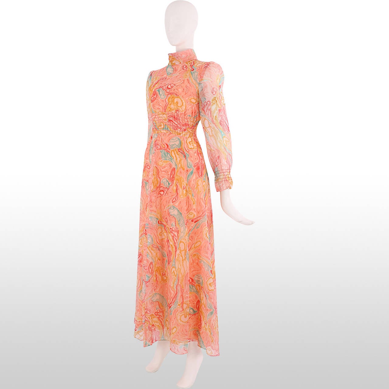 This 1970's gown is made from a printed chiffon overlay in tones of peach, turquoise, mustard yellow and crimson to create a marbled effect. It has a high standing collar and full length sleeves. The waist is nipped in with a wide band of elastic