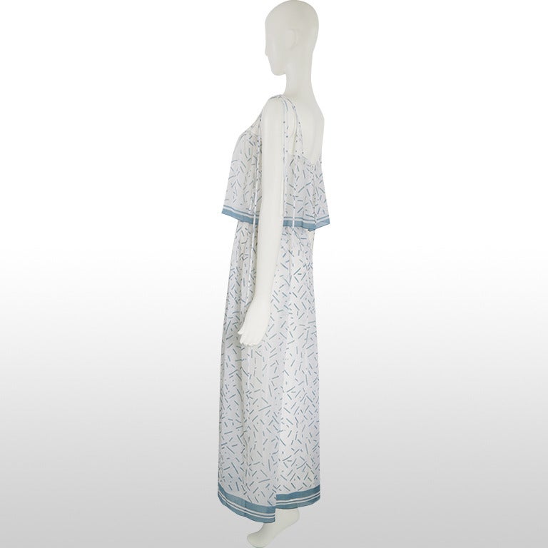 This gorgeous 1970’s summer dress has a caped overlay to the waist and a long skirt that gathers at the waist to create a figure flattering shape. It has strapped shoulders that tie and drape in strands. The print depicts short lines of blue on an