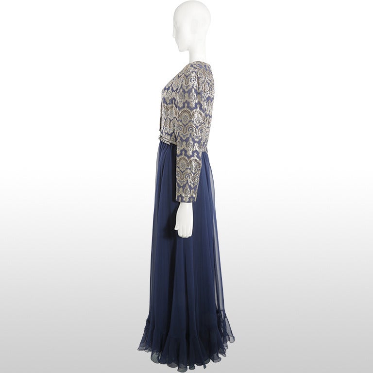 1970's navy gown with metallic brocade detailing on the bodice and sleeves. The belt is the same print as the embellished bodice. There are buttons from the neckline to the waist. There is a full chiffon skirt with a slight ruffle at the hem.