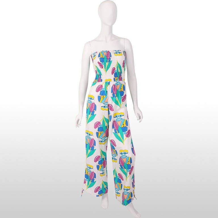 This fabulous and rare Fiorucci early 1980's jumpsuit has a comical print of clowns and tulips in bright colours such as blues, purple, yellow and green and is printed on white cotton fabric. It has an elasticated smocked strapless bodice and flared