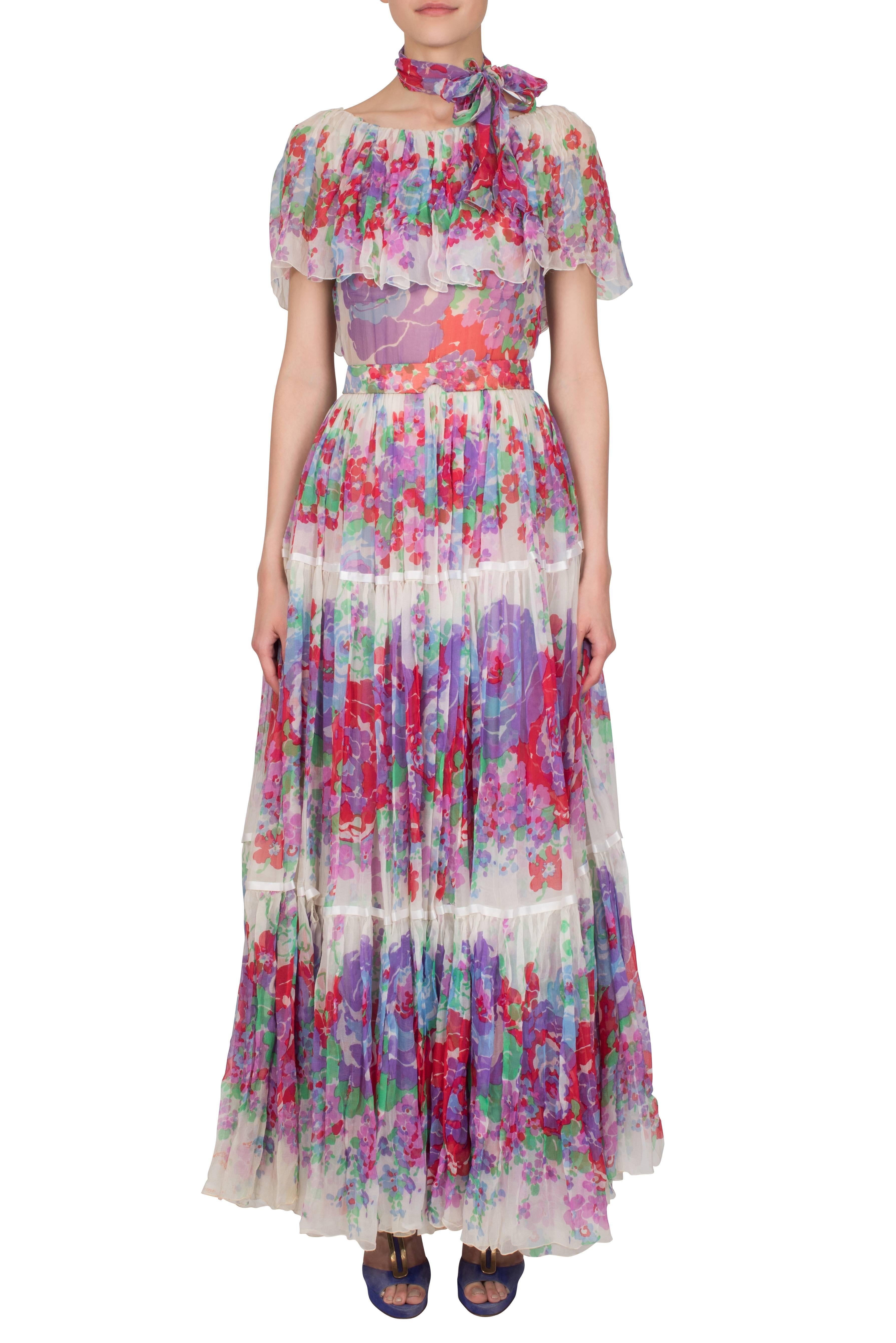 A sheer ivory chiffon maxi dress by London couturier Harald which features a delicate lilac, pink, red, blue and green watercolour floral print. The dress features a pleated and tiered skirt divided by two white satin ribbon trims; the lower trim