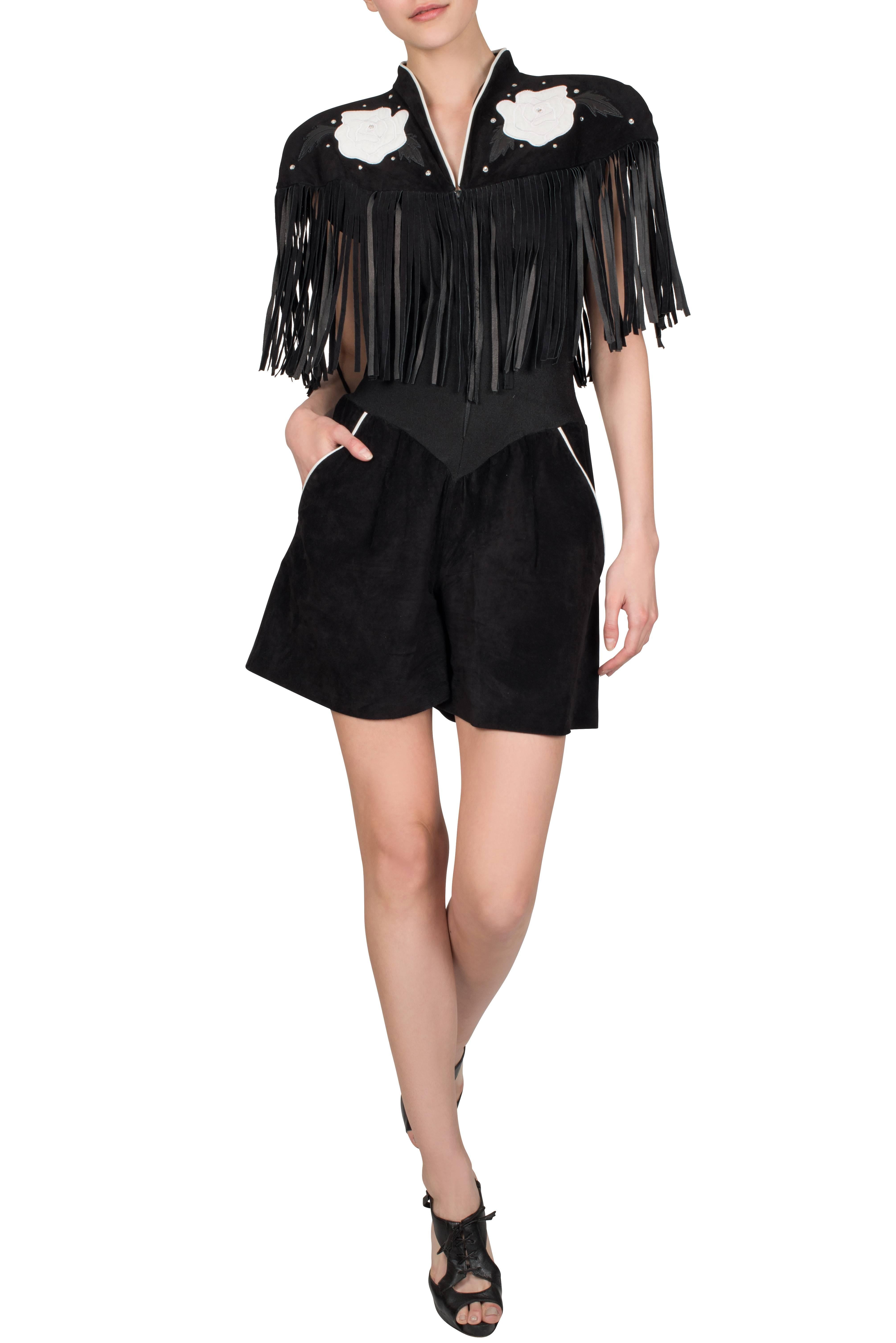 A 1980's Caché black suede romper, with cowboy style fringes circling the upper part of the top, cap sleeves and back. White leather rose appliqués and rhinestones embellish the front and centre back. The centre part of the bodice is made of a