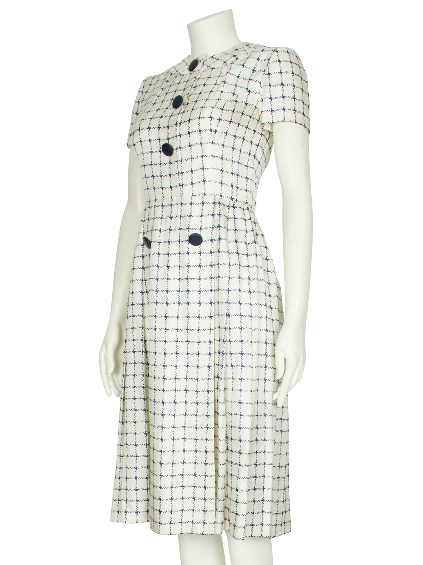 A short-sleeved ivory silk day dress by London society couturier Harald which features an all-over navy blue checked print and oversized navy blue buttons. The soft, lightweight dress is straight cut to the waist and features a round neckline with a