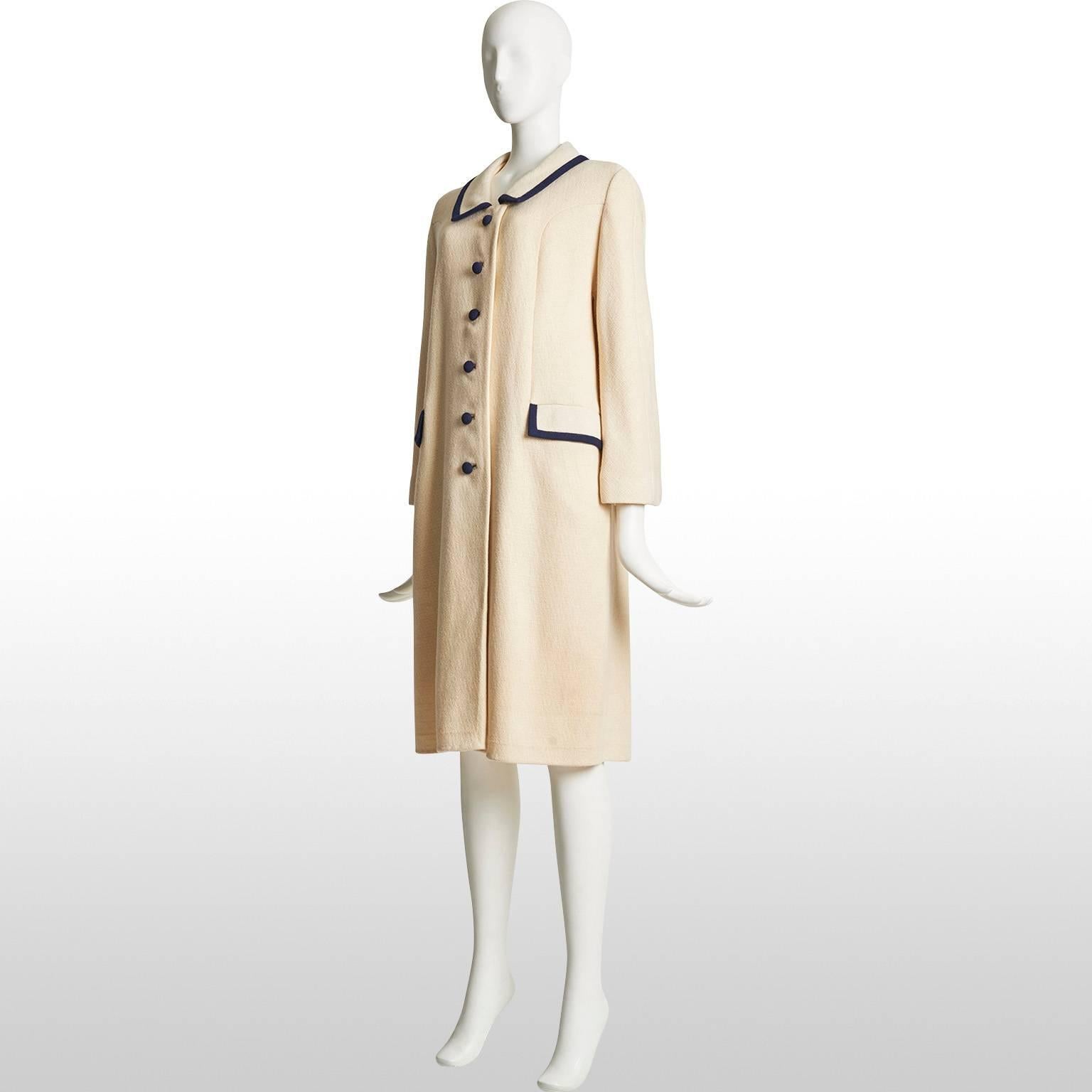 This is a ivory wool mid length coat with navy trimming from the 60s. The coat is single breasted with six navy blue buttons, the side pockets are horizontally aligned and there is a half belt on the back side of the coat. The piece remains in