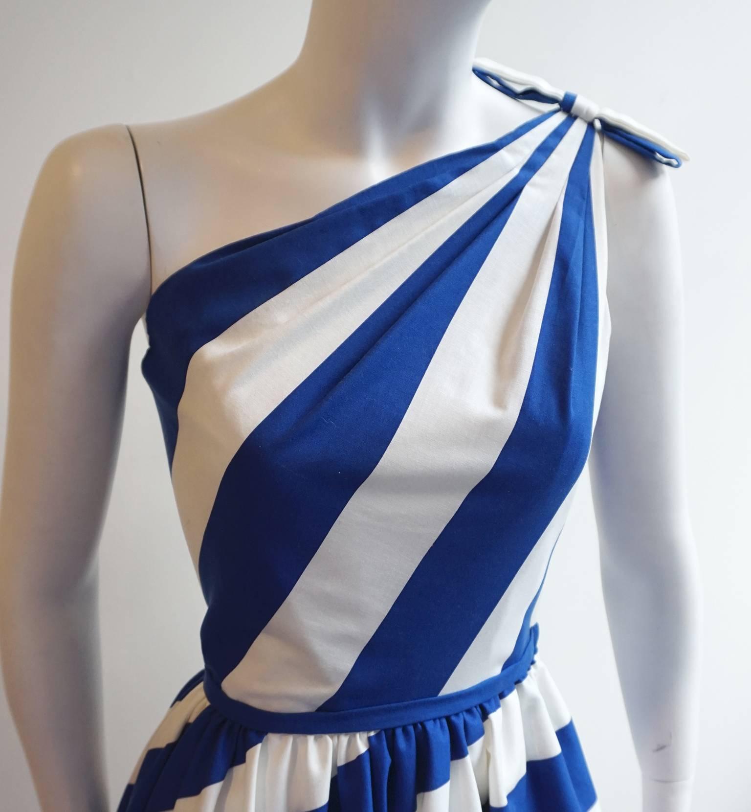 This VICTOR COSTA dress is made of a light weight cotton. The one shoulder bodice features a bold blue and white stripe originating at the top of the shoulder, where a matching blue and white bow sits. There is piping along the waist line. The skirt