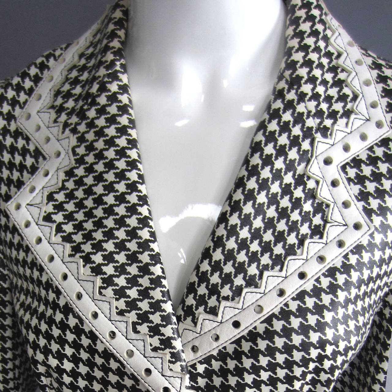 This jacket is made of leather that is covered in a black and white houndstooth print. The printed body is accented with white, pointed trim. The trim is featured on the collar, along the front opening, and along the hem. The front features 3 large,