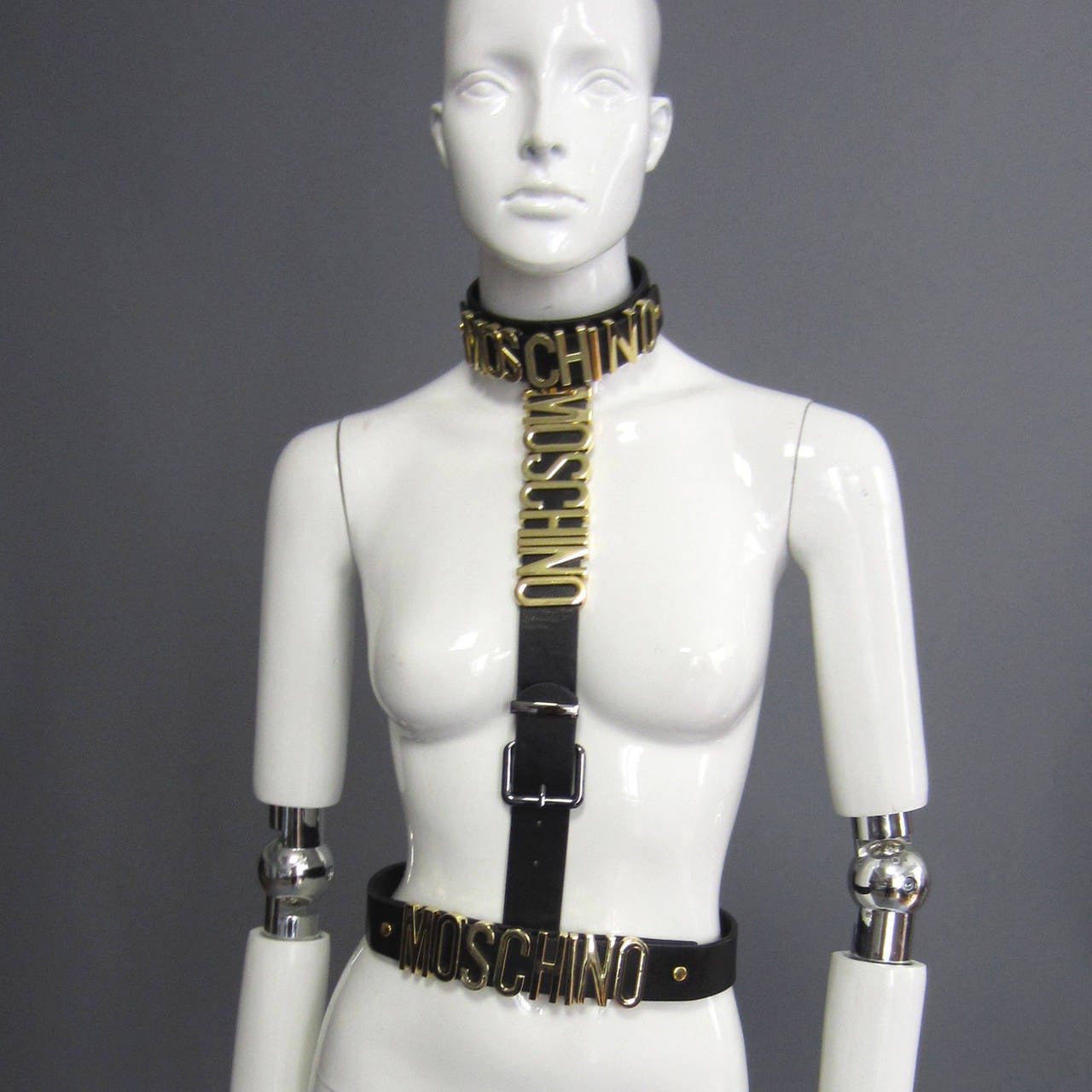 MOSCHINO, MOSCHINO, MOSCHINO! This leather accessory with gold hardware says it 3 times! This piece features a choker and belt, both with the MOSCHINO logo. These two pieces are connected through snaps with a center harness piece, that also