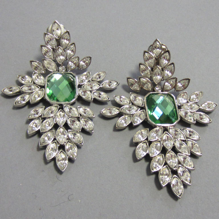 SCAASI stament earrings. These earrings feature a green center stone. The articulated, leaf shaped rhinestones surround the center in a cross design. The top, bottom, and either side each feature a collection of these rhinestones configured in a