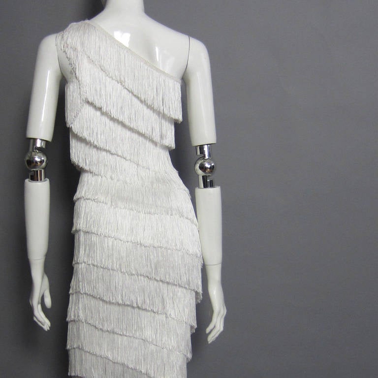 Rows of white fringe make up this striking dress from the genius herself, NORMA KAMALI. The one shoulder design is balanced by the asymmetrical hem. The fringe is staggered on a slight diagonal to match the asymmetry of the design. The lining is