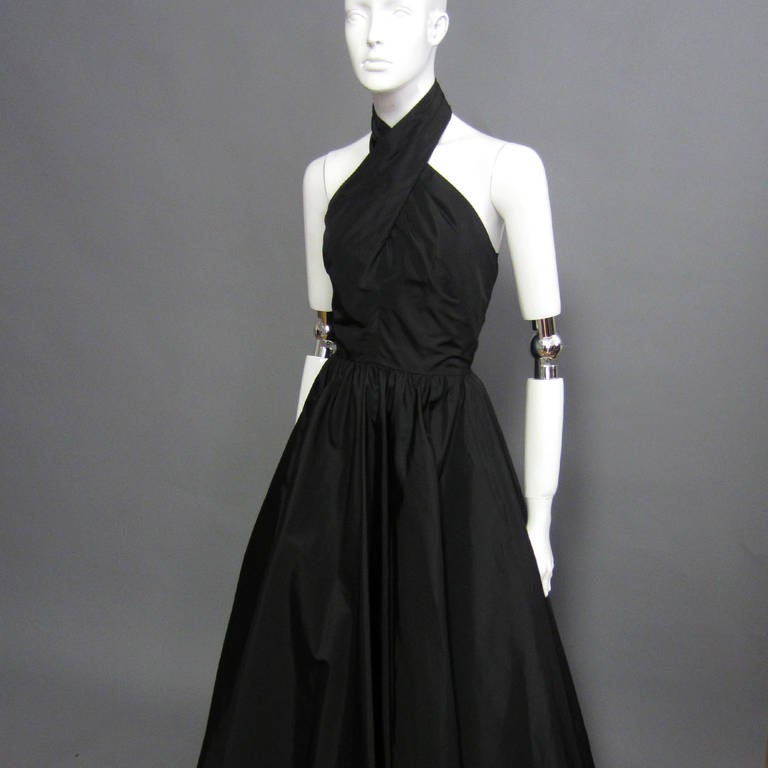 A 1980s take on Audrey Hepburn, this black Victor Costa dress is modern elegance with flair. The halter top is comprised of two, criss crossing straps that then wrap around the neck. They are secured at the back of the neck with a double hook and