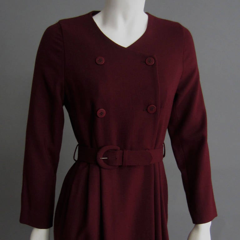 The rich burgundy color of this wool dress is perfect for fall. The dress is is designed to resemble a coat; it has a double breasted closure and matching fabric belt. There is slight pleating at the waist that conceals the side pocket details of