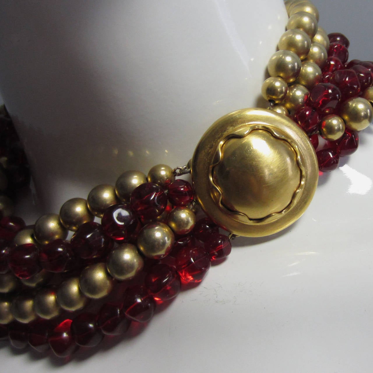 This necklace features 3 strands of red glass beads and 3 strands of gold beads. The strands intertwine when worn, creating a layered effect. There is a gold, articulated circe clasp closure that can be worn to the back, side or front as a pendant.