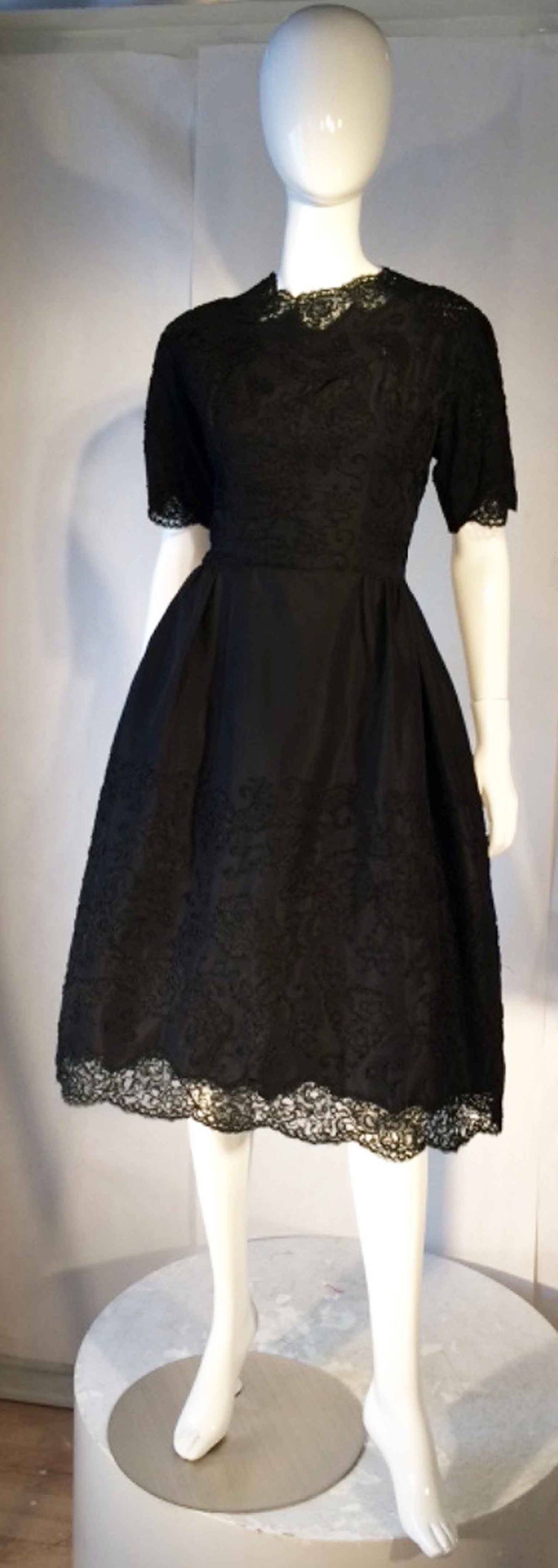 A fine and rare Sophie Gimbel couture cocktail dress. Hand constructed black silk faille item features matching black applique lace trim and hem. Item fully lined with a cinched waistline, boned bodice with metal zipper up back. Numbered Saks Fifth