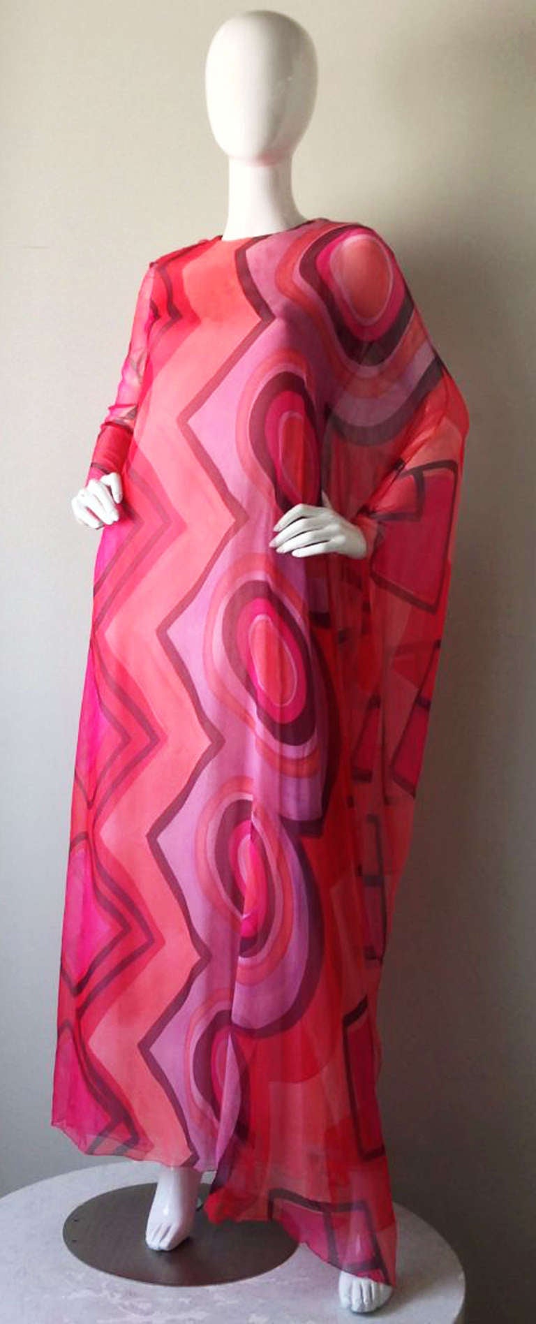A stunning Gagliano chiffon gown. Contrasting pinks graphic print chiffon item fully silk lined and zips up back. Gown features attached flowing caped single sleeve and opposite full sleeve. Pristine, appears unworn.
