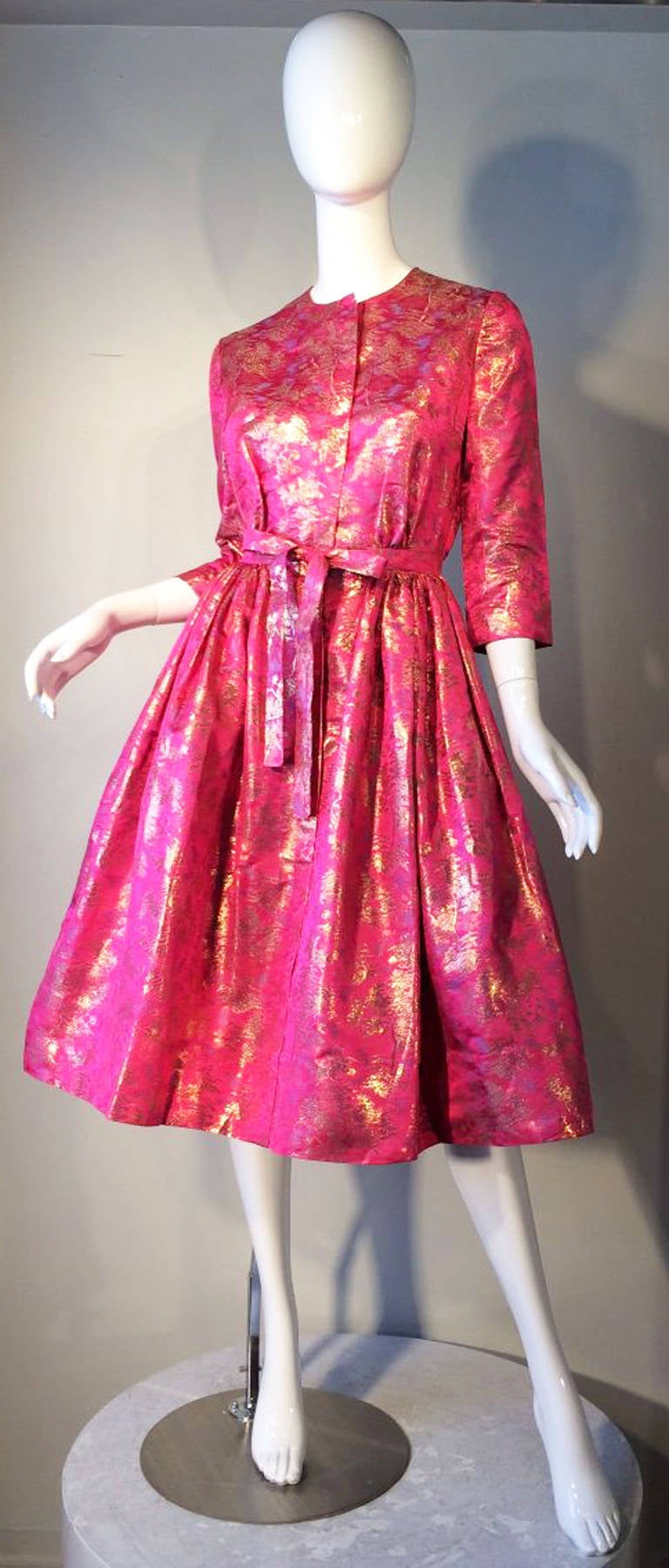 A fine vintage Larry Aldrich cocktail dress. Vivid pink silk brocade fabric features woven gold metallic details. Fully silk lined item features a nipped waist and full pleated sides skirt. Hidden button front closures with a attached bow tie front