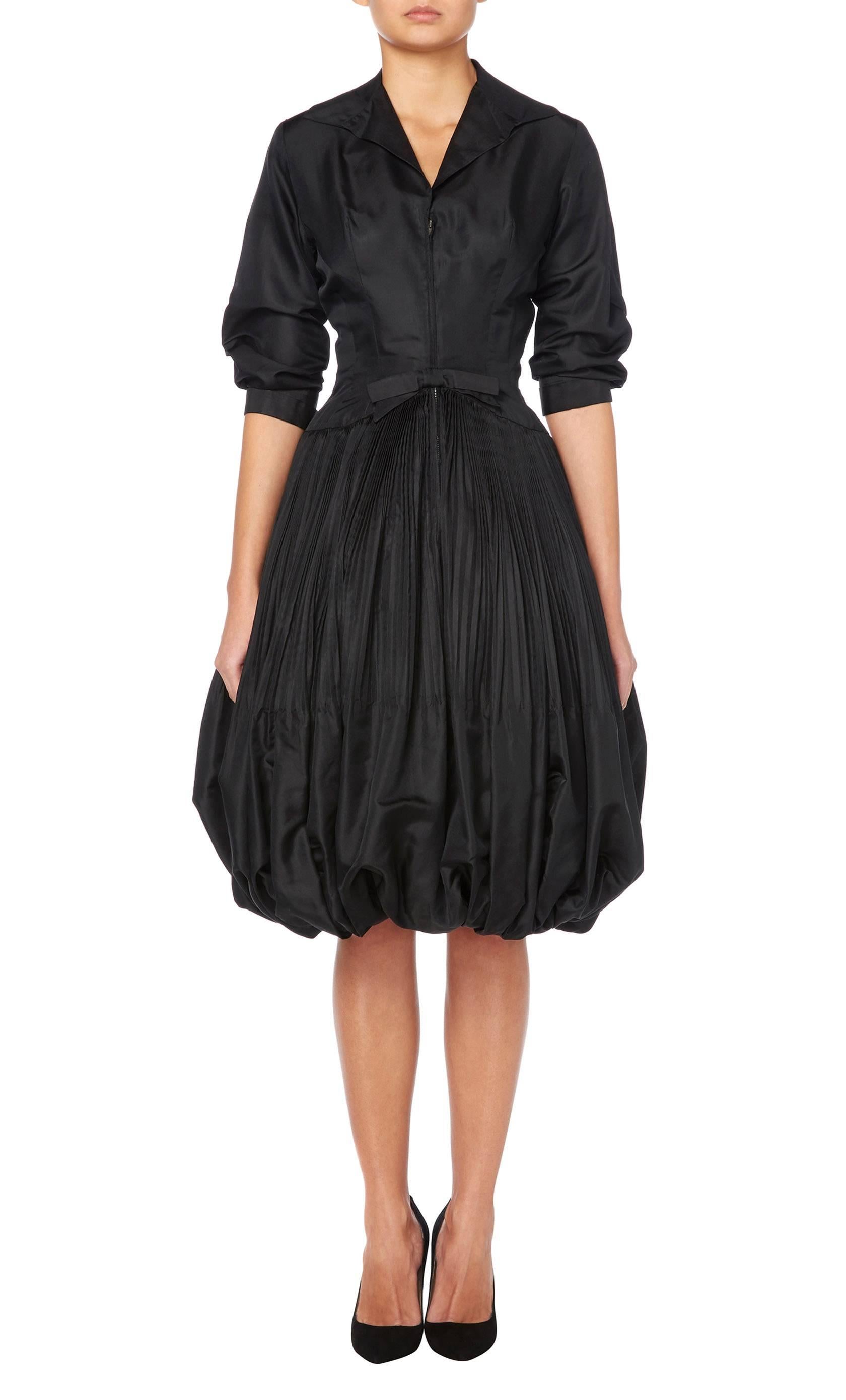 A wonderful LBD by Seymour Jacobson, this cocktail dress features a collar, three-quarter length sleeves and a full puff balled skirt with micro-pleating. A zip fastens to the front with a bow detail on the waist, making this beautifully tailored