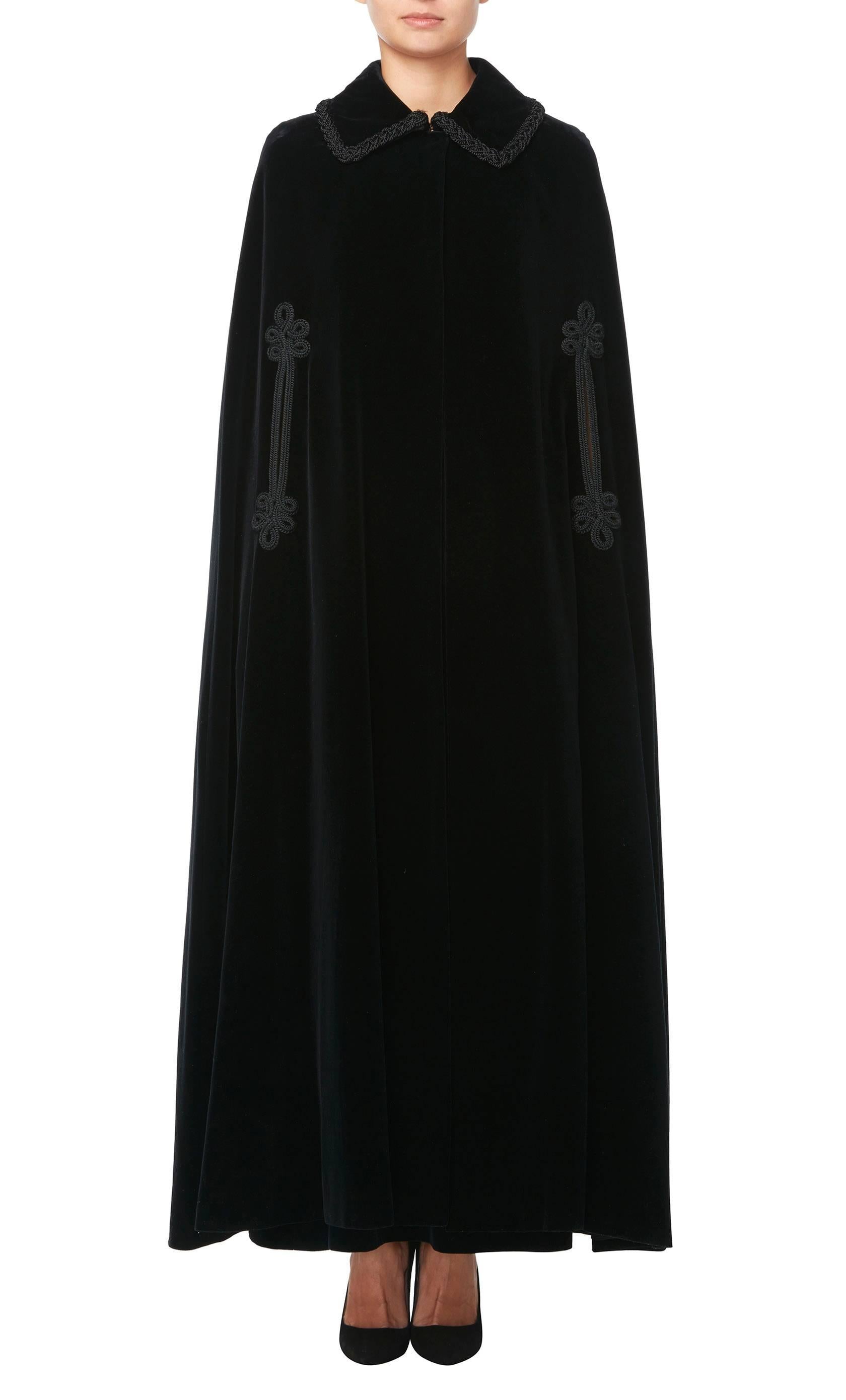 A dramatic evening ensemble, this Jean Patou two-piece is made up of a full-length cape and maxi skirt in matching black velvet. The cape features a collar and a single hook and eye fastening at the neck, allowing a glimpse of the outfit beneath.