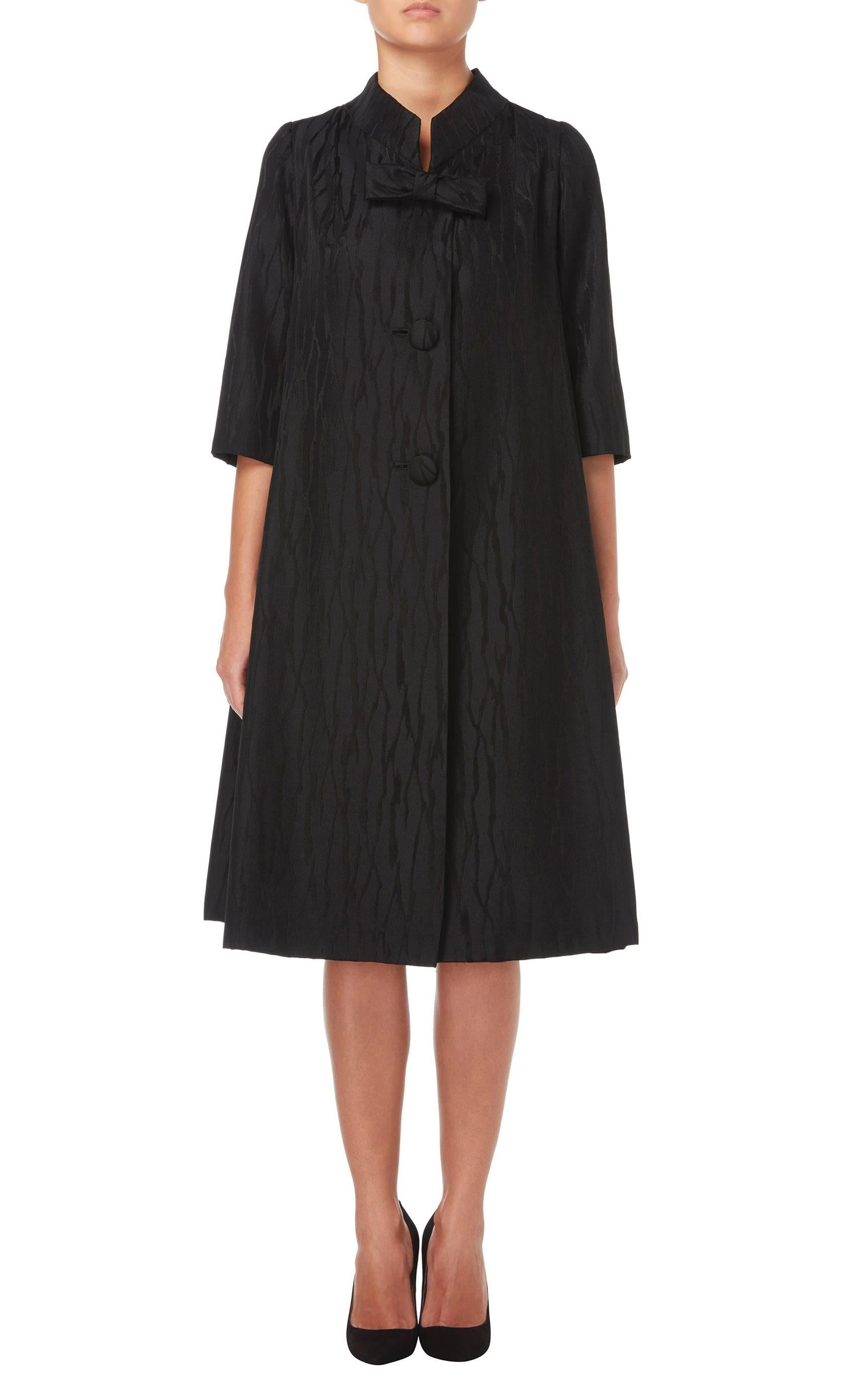 This chic swing coat in jacquard black silk is a fabulous option for an evening event. The A-line cut, wide standing collar and bracelet length sleeves create a classic silhouette, while the large fabric matched buttons and bow on the bust add a