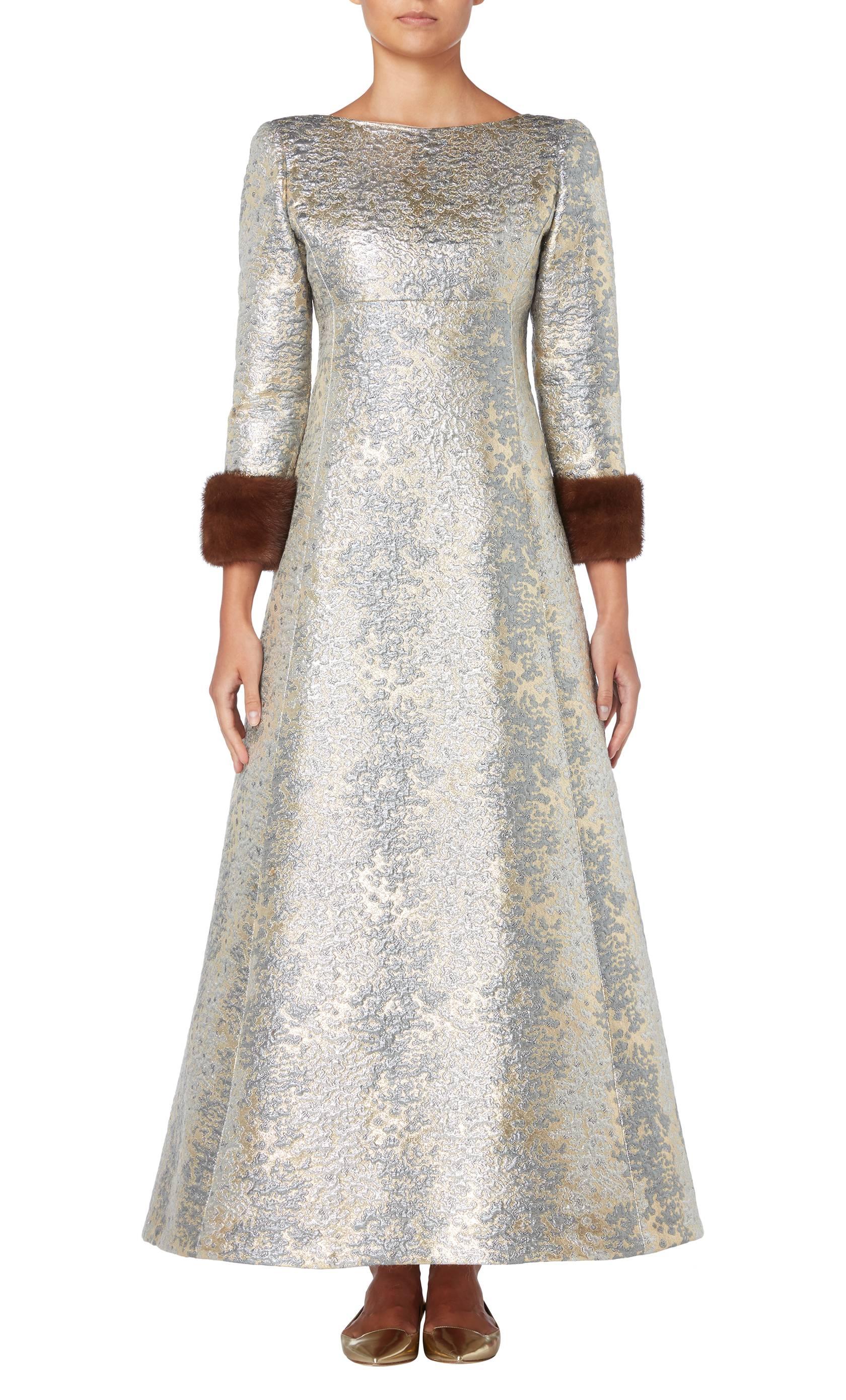 This Malcolm Starr evening dress will make an opulent choice for red carpet events. Designed by Elinor Simmons, the dress is constructed from blue, silver and gold brocade and features three-quarter length sleeves with brown mink fur cuffs. A square