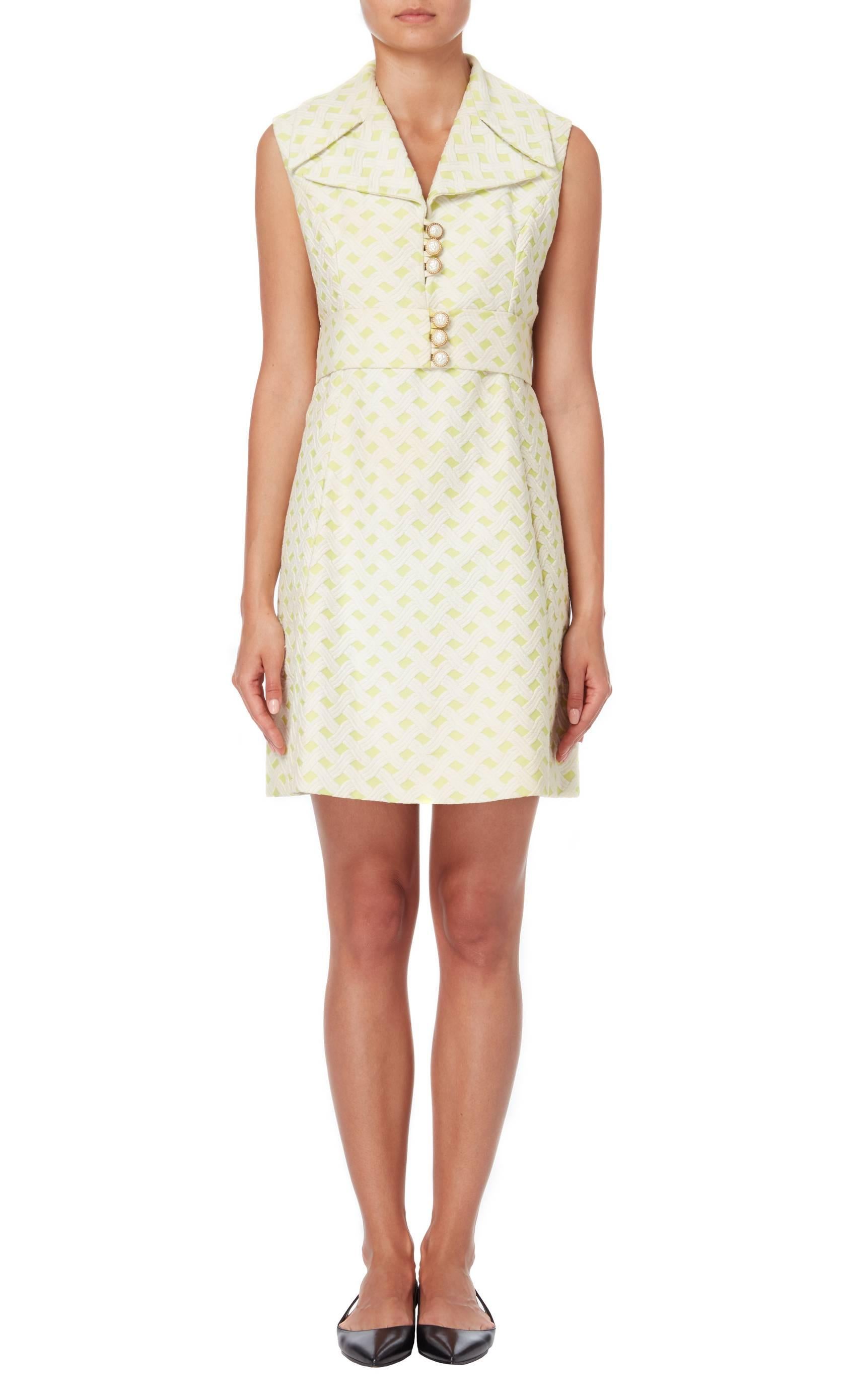 Constructed in a cotton and synthetic mixed fabric, the material of the dress is made in a jacquard hatch cross pattern of pale green and white. Large pearl and gold buttons adorn the bodice and waist belt, with large oversized collar and lapels.