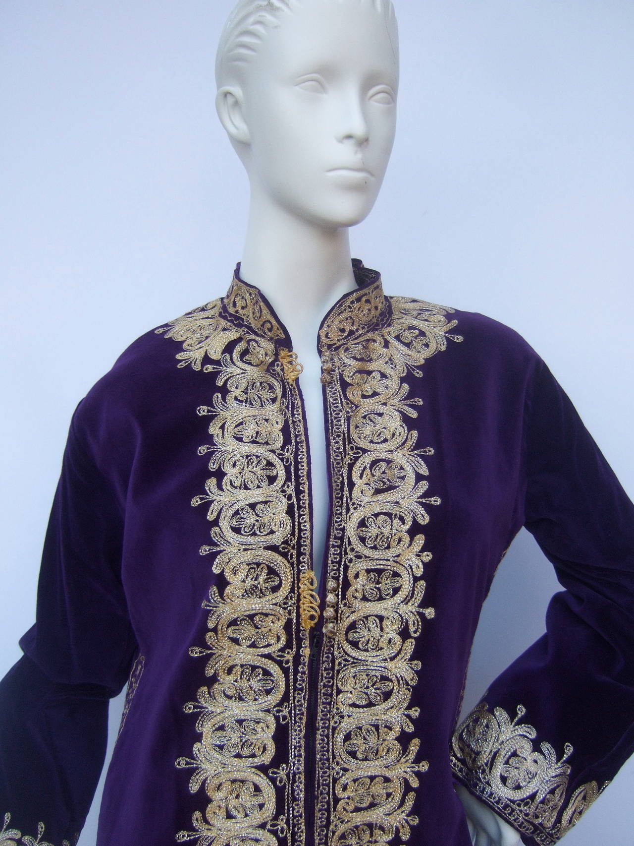 Exotic violet velvet gold embroidered evening caftan c 1960s
The Moroccan style bohemian caftan is designed with regal purple velvet
Embellished with ornate gold metallic embroidery that circles the neckline, runs down the center zipper, extends