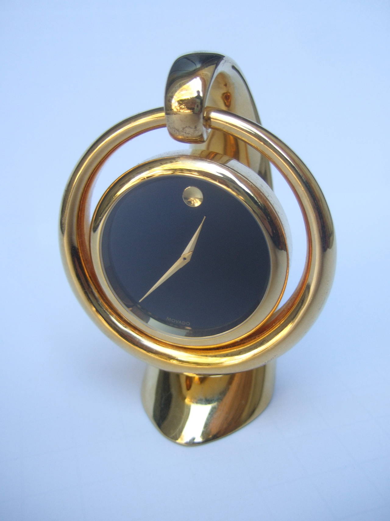 Movado sleek gilt metal diminutive desk clock 
The elegant clock is designed with Movado's signature face dial
The compact circular clock mechanism is suspended from the curved base
The iconic black face dial is stamped Movado 

The stylish