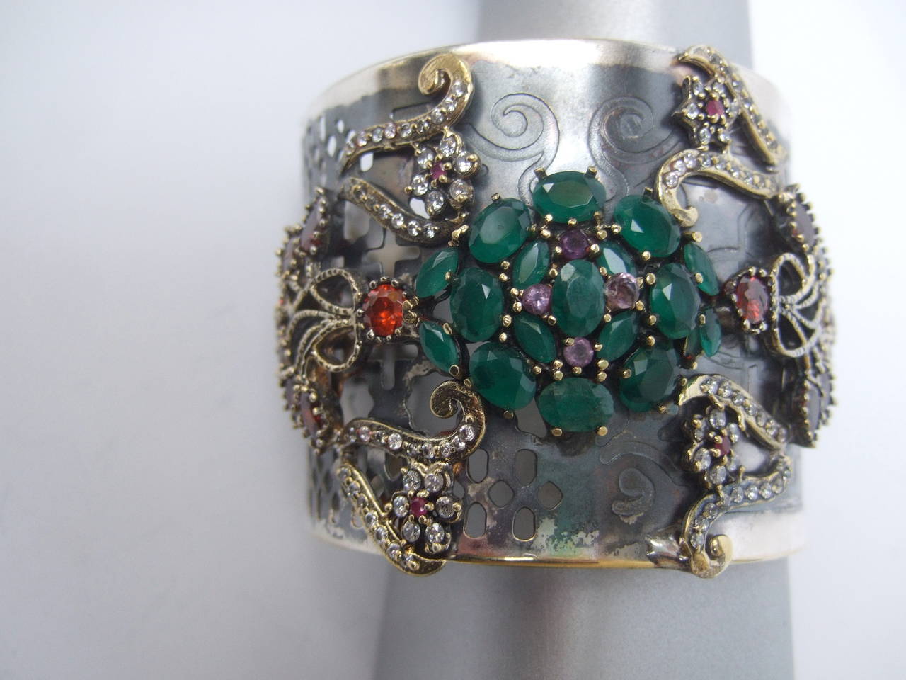 Crystal jewel encrusted wide silver metal cuff bracelet
The exotic cuff is embellished with a cluster of emerald green
glittering crystals in the center accented with four tiny amethyst  
crystals. The sides are enhanced with sinuous rows of